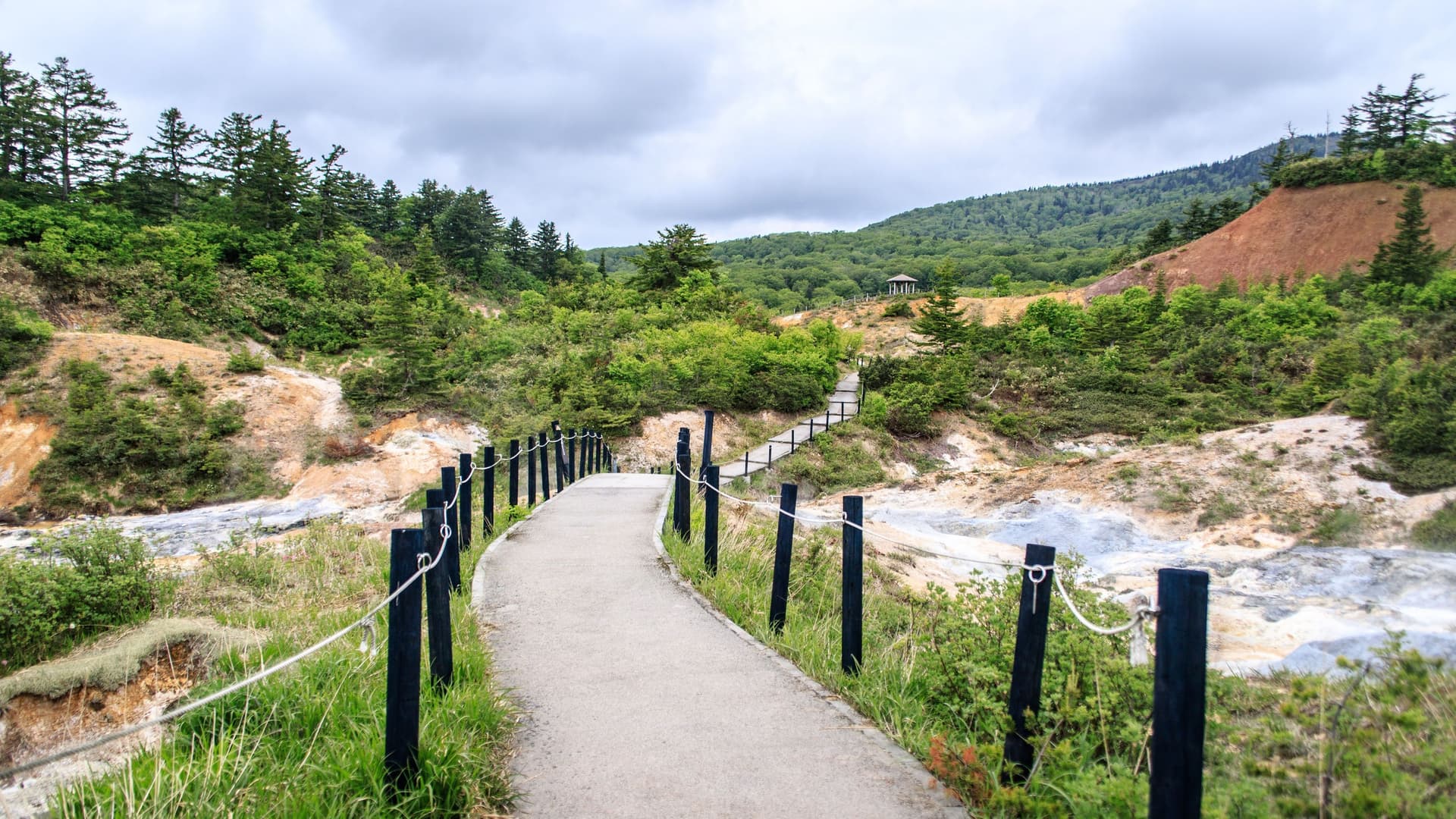 A winding elevated walkway with a rope fence leads through the volcanic landscape of Akita, Japan, with green trees and a small gazebo visible in the distance.