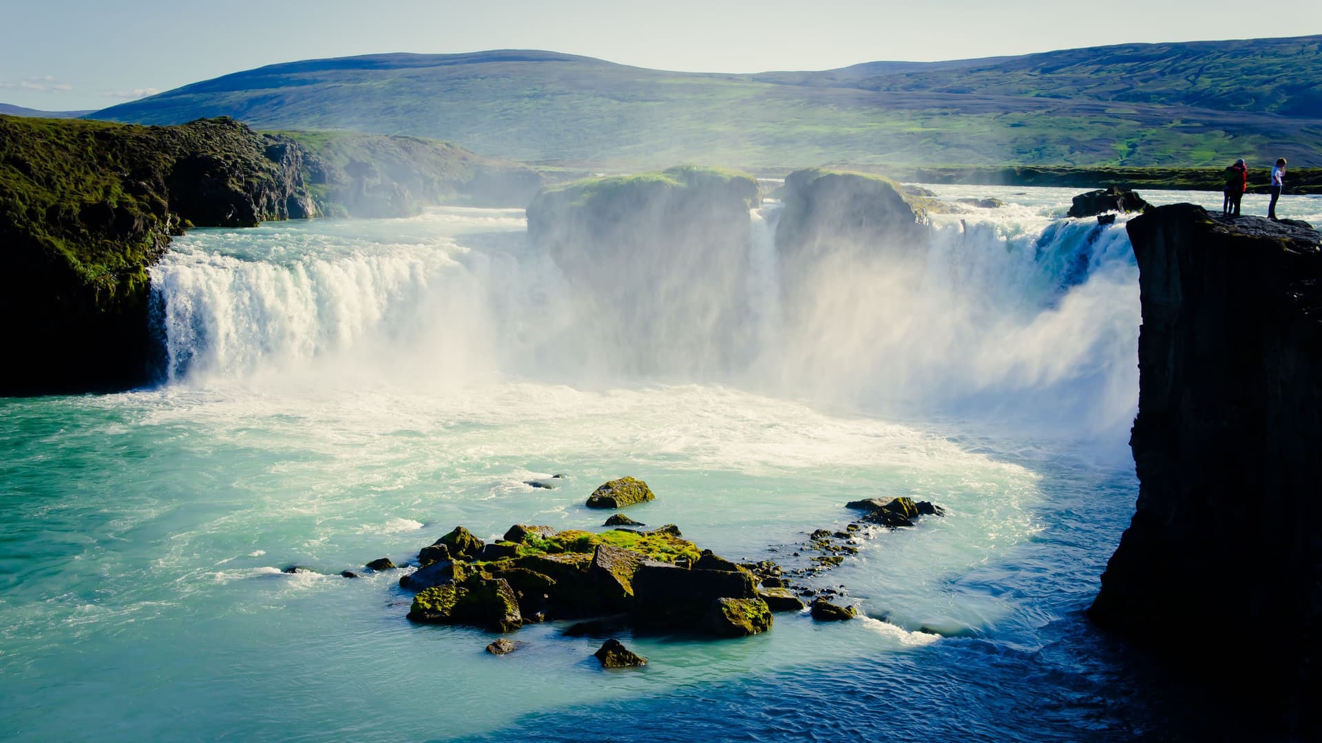 A breathtaking view of Goðafoss, the "Waterfall of the Gods," in northern Iceland, with powerful cascades of turquoise water and two people standing on the rocky cliff.