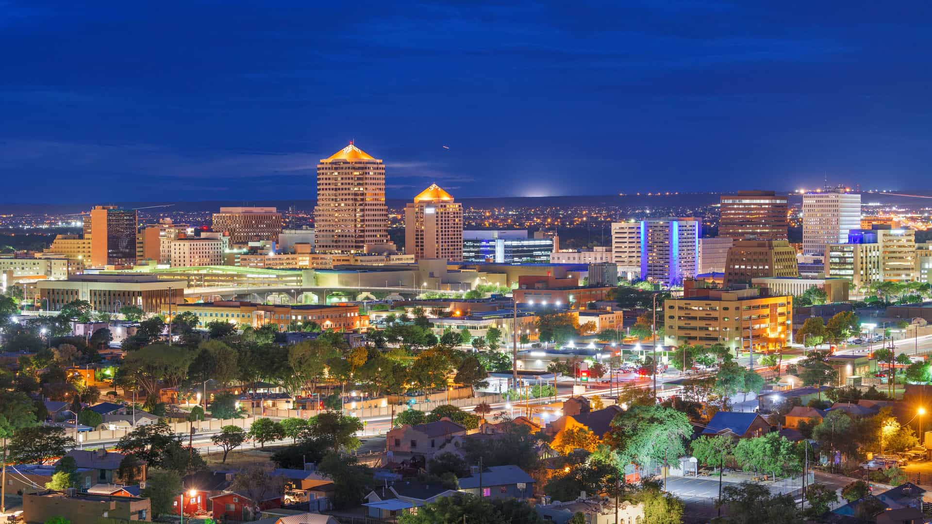 A panoramic nighttime view of the downtown skyline of Albuquerque, New Mexico, with its many buildings and streets lit up. The city glows under a deep blue twilight sky, with light trails from cars visible on the streets in the foreground.