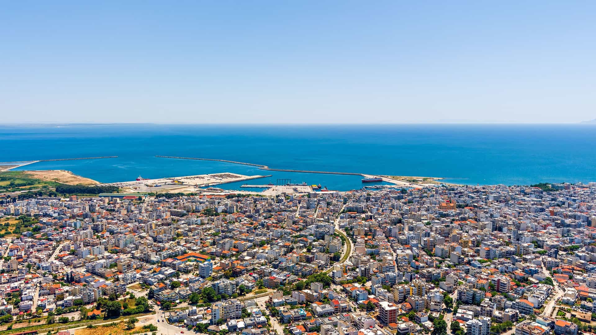 An aerial view of the coastal city of Alexandroupolis, Greece. Densely packed white buildings with red roofs extend to a large, busy port, where a long breakwater and piers jut into the deep blue Aegean Sea. The sky is clear and bright blue, stretching to meet the sea at the horizon.