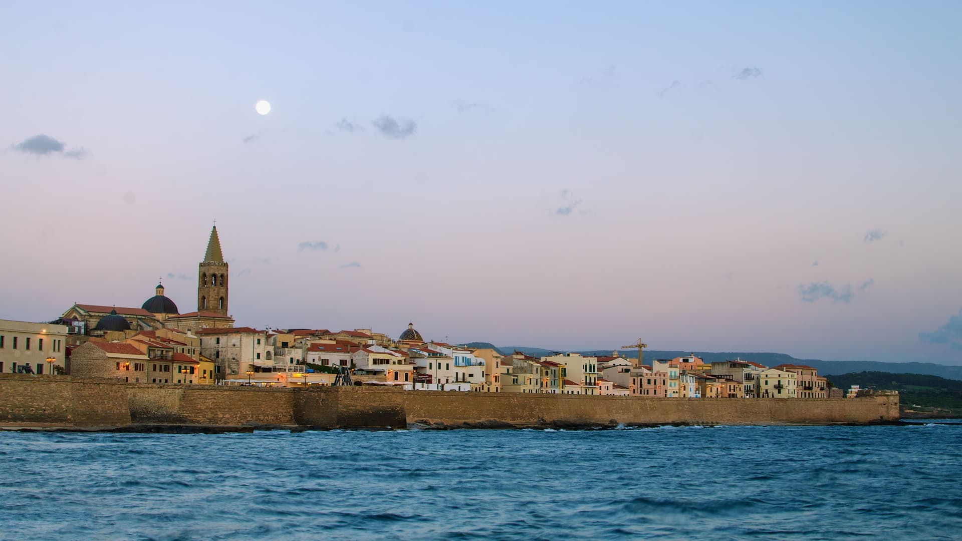 A panoramic twilight shot of Alghero, Italy, with its historic city walls and a bell tower silhouetted against a pastel sky with a full moon, reflecting on the Ligurian Sea.