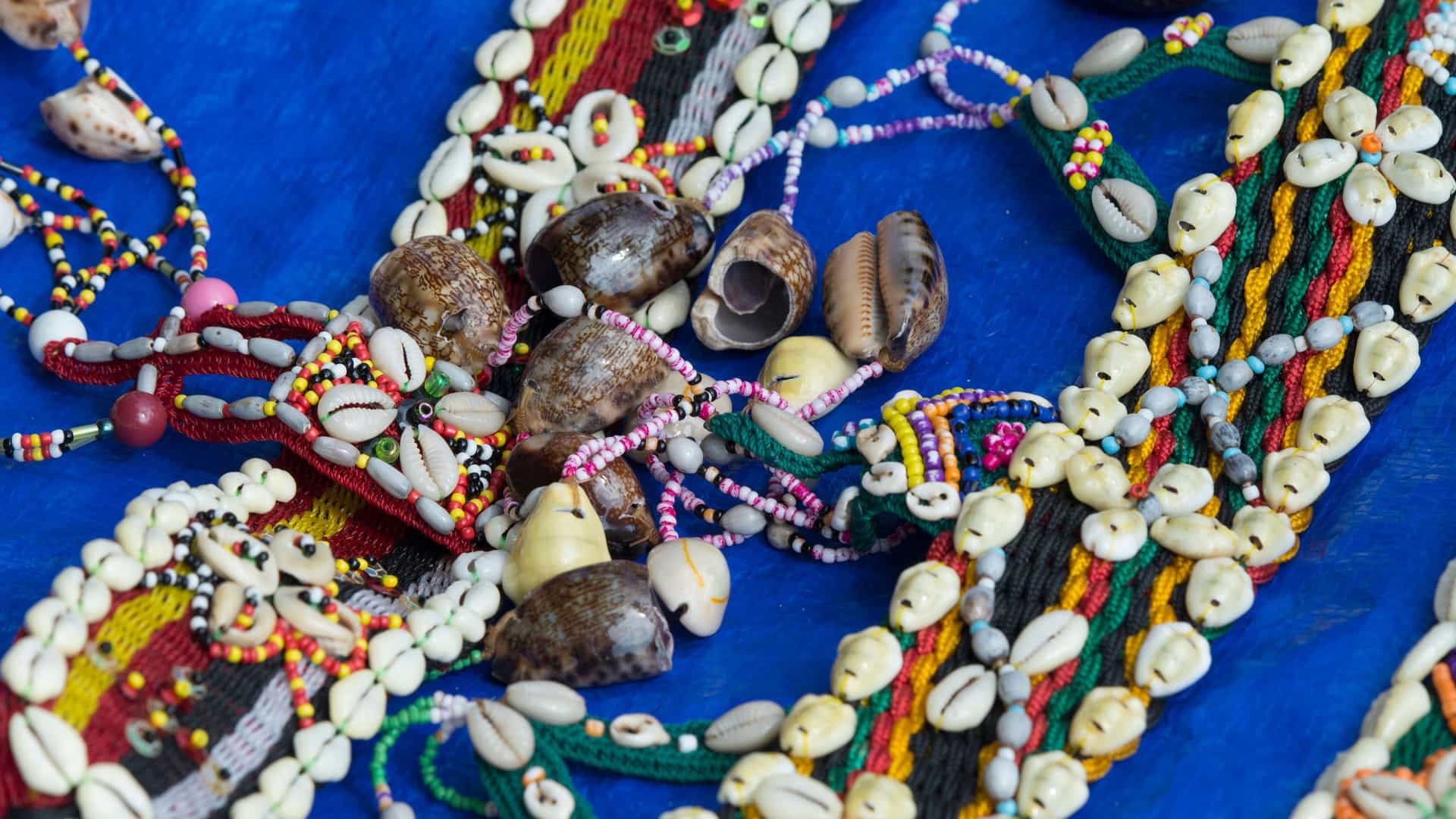 A close-up of a variety of handcrafted jewelry, including necklaces and bracelets made from cowrie shells and colorful beads, laid out on a blue cloth.
