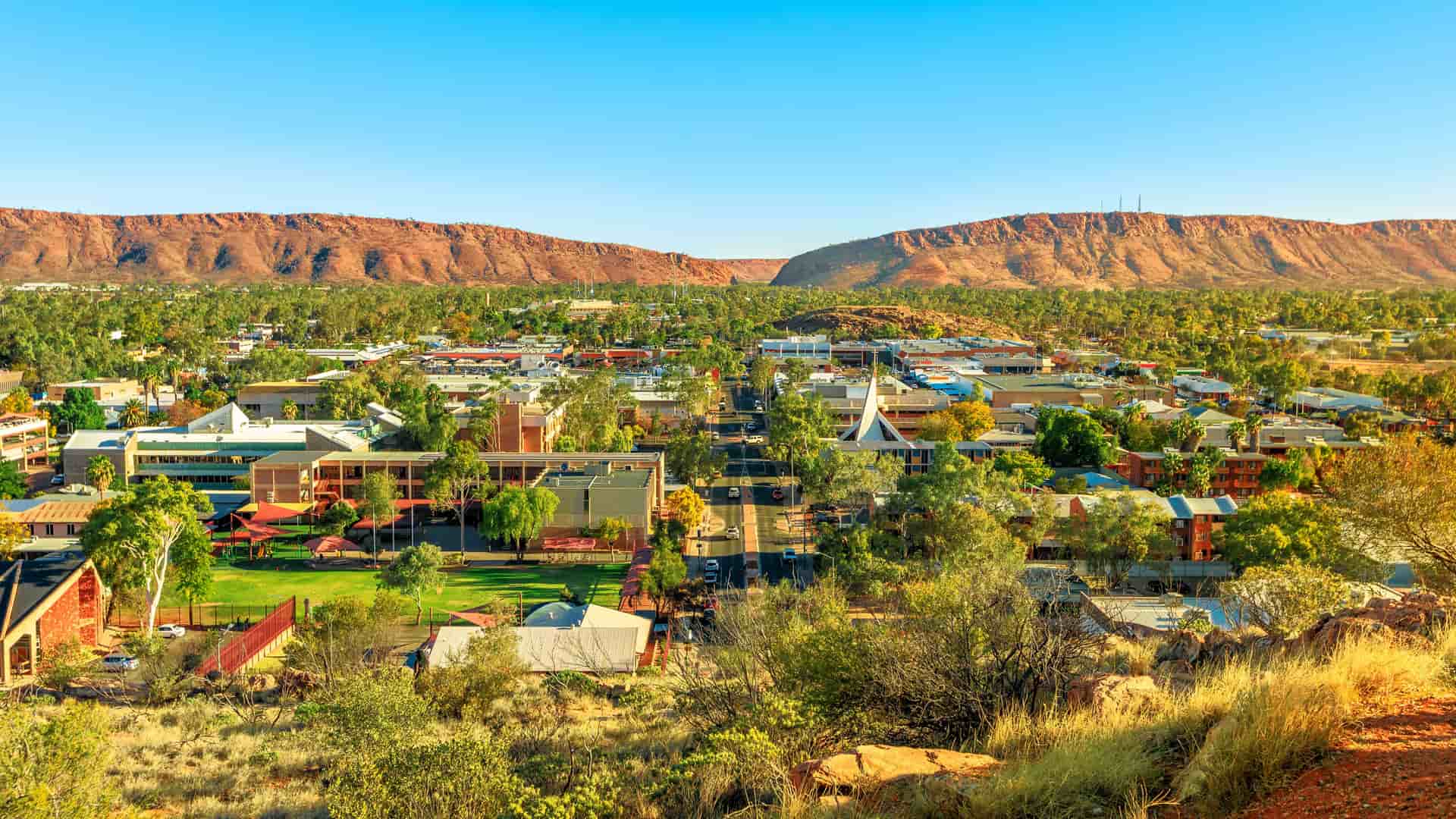 A panoramic, elevated view of the town of Alice Springs, Australia, nestled in a valley between two large, rugged red-rock mountain ranges. The low-rise buildings and green parks of the town are bathed in a warm, golden light under a clear blue sky, contrasting with the arid desert landscape.