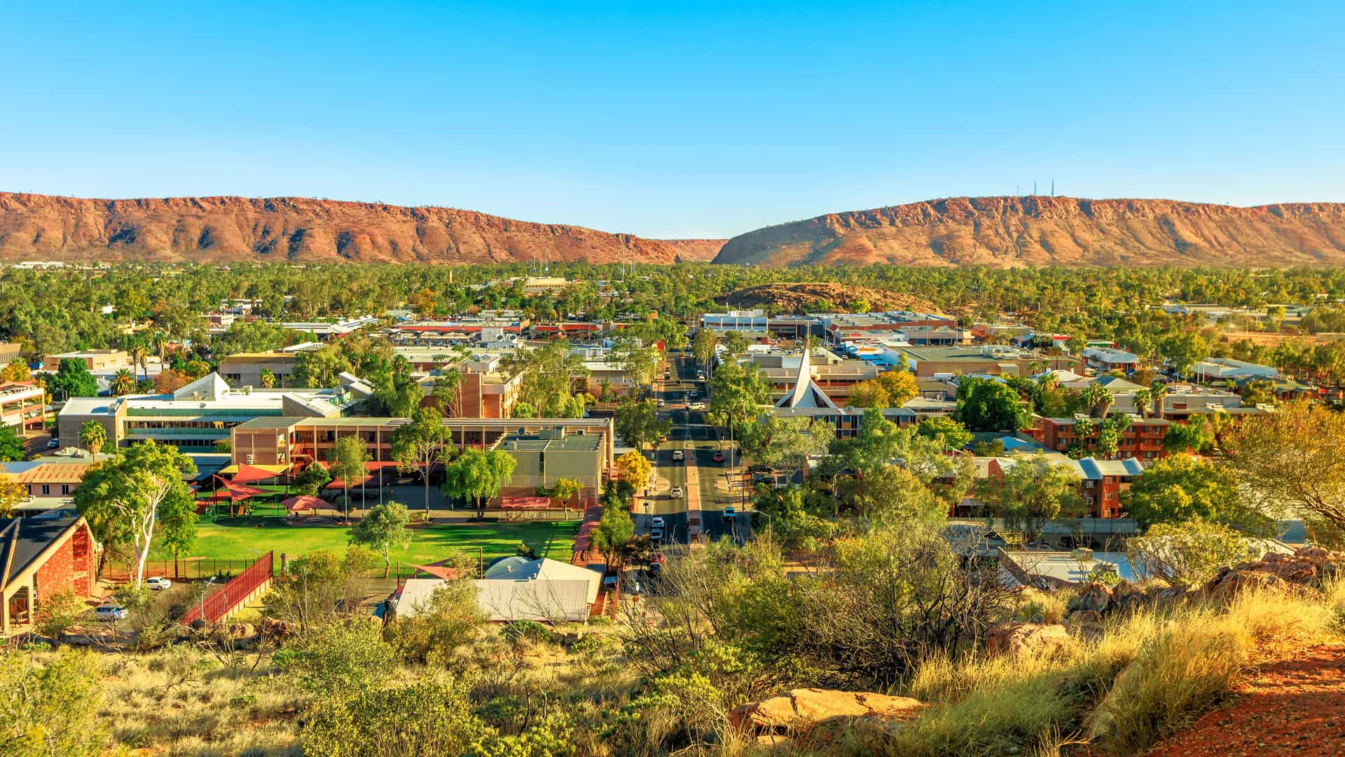 A panoramic, elevated view of the town of Alice Springs, Australia, nestled in a valley between two large, rugged red-rock mountain ranges. The low-rise buildings and green parks of the town are bathed in a warm, golden light under a clear blue sky, contrasting with the arid desert landscape.