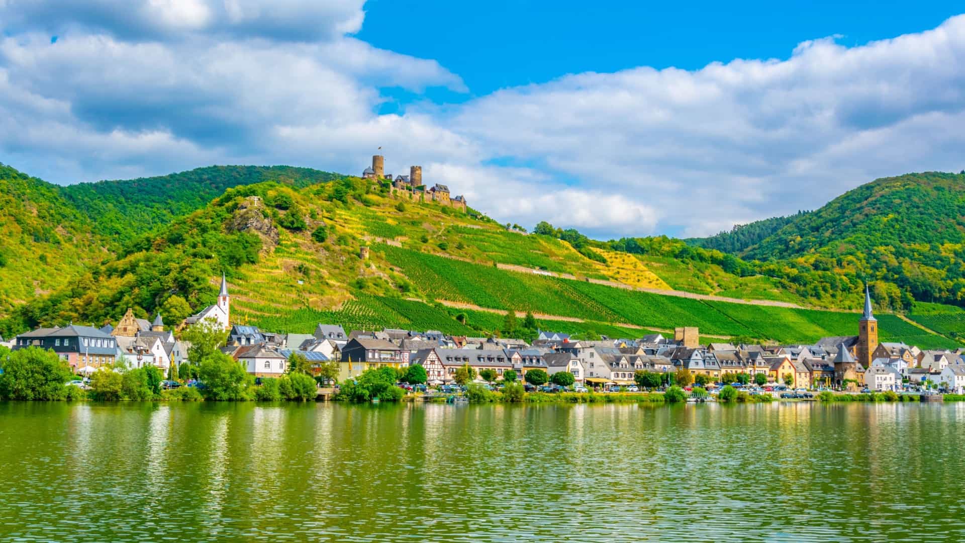 A beautiful landscape of Alken, Germany, showing a picturesque village nestled on the banks of the Moselle River, with vineyards and the Thurant Castle on the hill above.