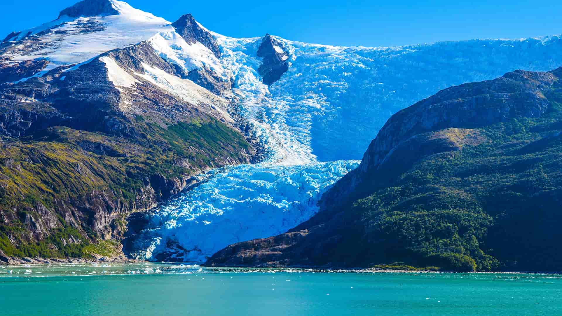 A majestic view of Alley Glacier, a massive, flowing blue glacier, nestled between two steep, rocky mountains that meet the calm, turquoise water.