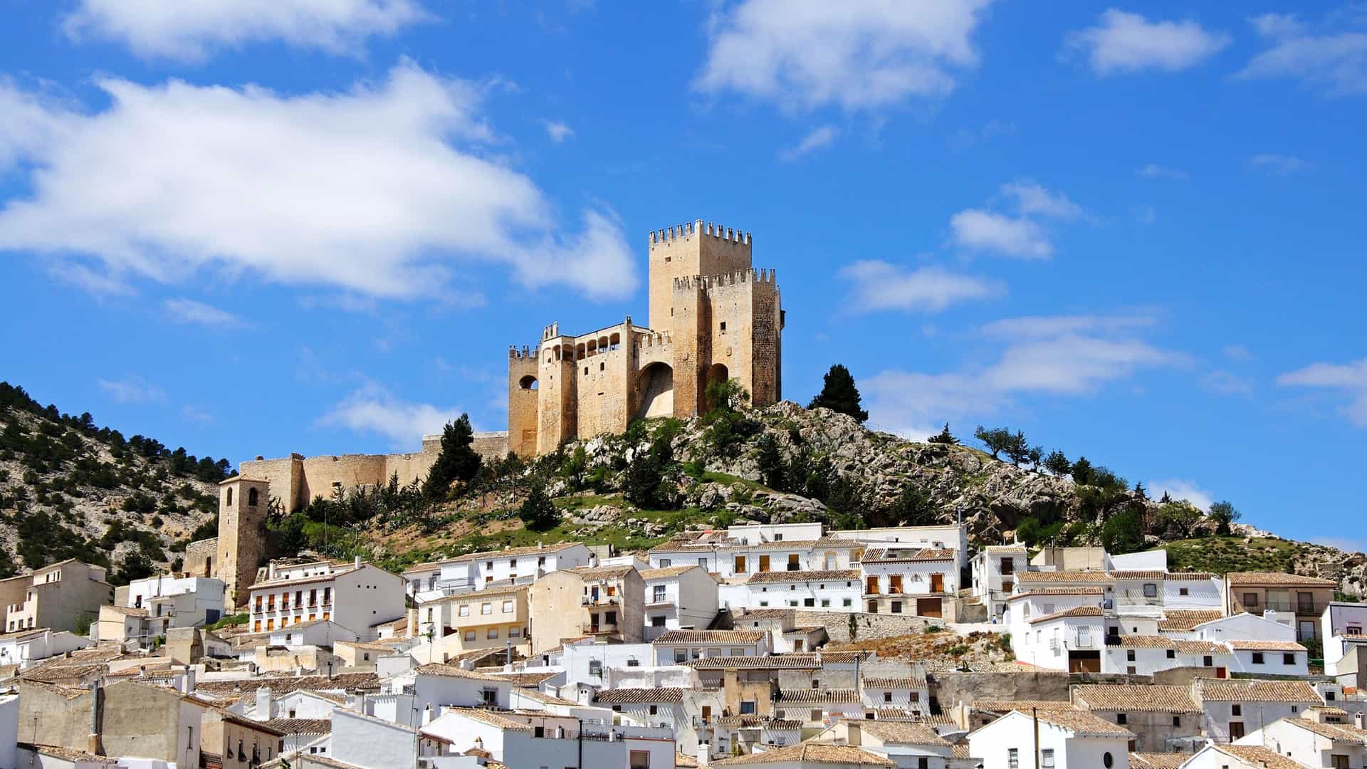 A historic fortress, the Castle of La Calahorra, stands on a hilltop overlooking a town with white-washed houses below, under a blue sky with scattered clouds in Almería, Spain.