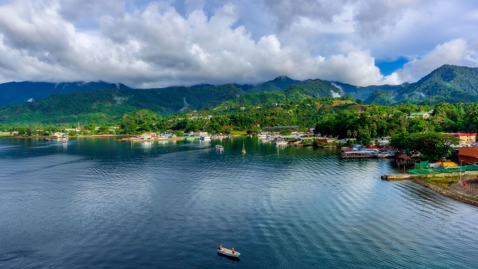 A scenic view of Alotau, Papua New Guinea, with a vibrant harbor and a small town nestled against a backdrop of lush green mountains under a cloudy sky.