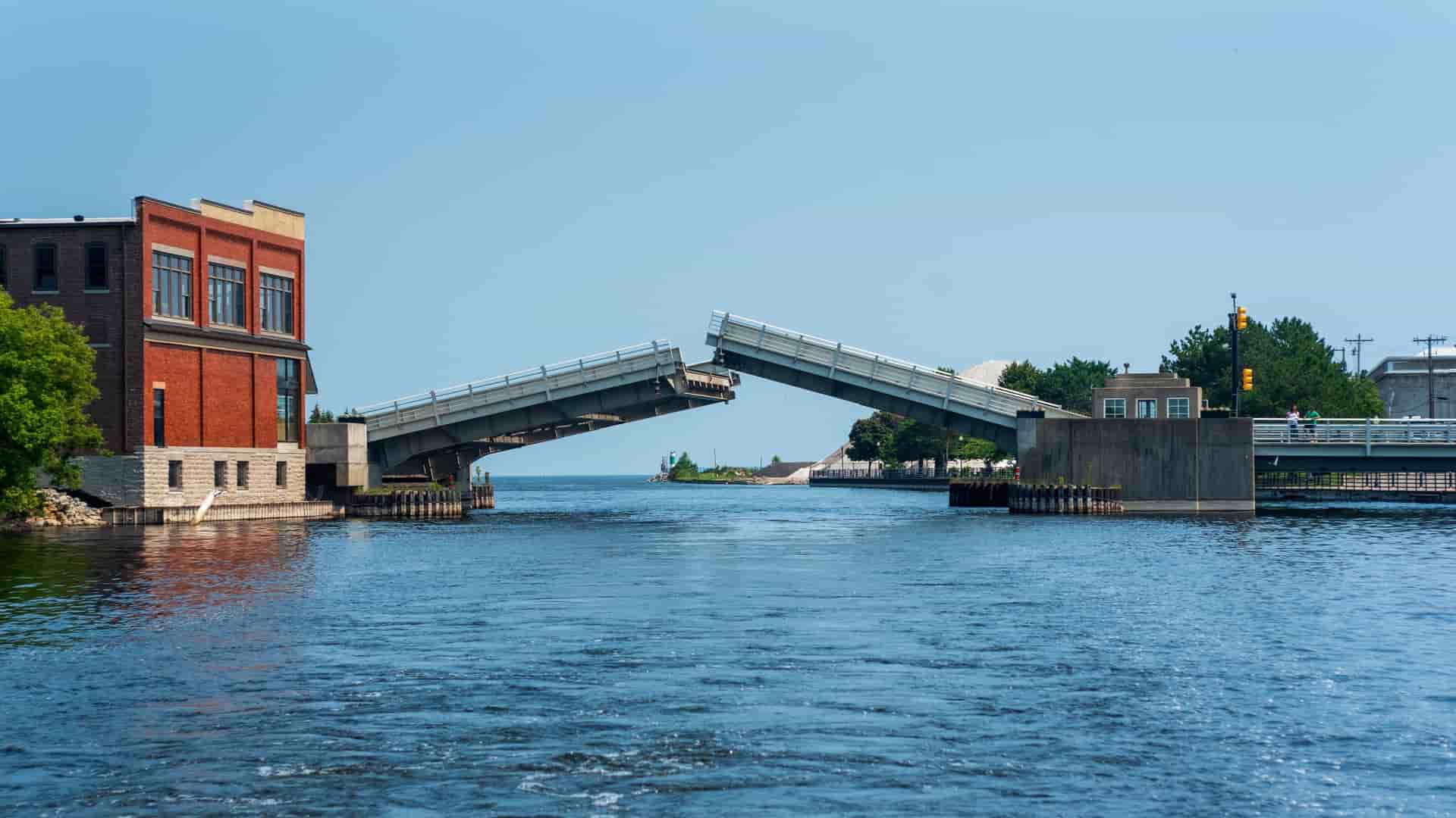 A drawbridge over a river in Alpena, Michigan, is shown in a raised position, with a red brick building on the left bank and a small park on the right.