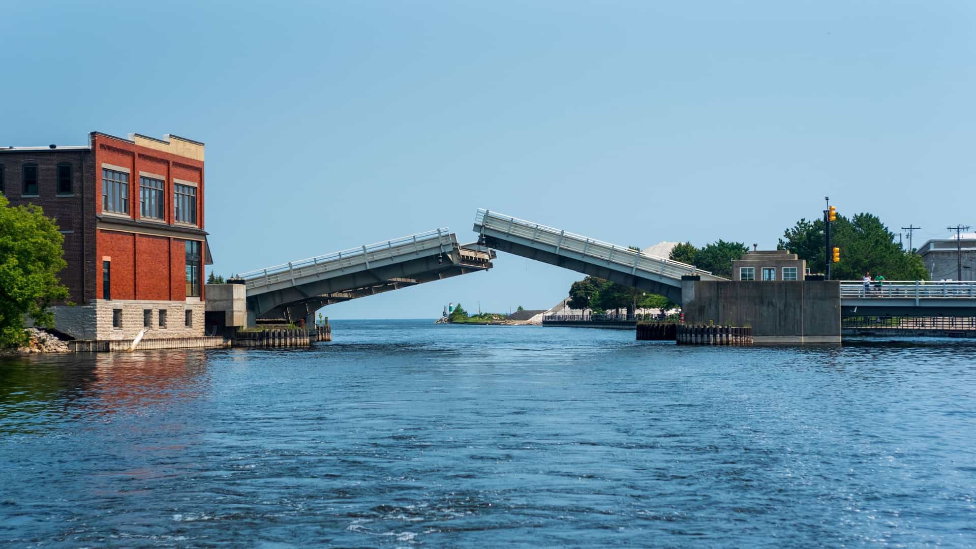A drawbridge over a river in Alpena, Michigan, is shown in a raised position, with a red brick building on the left bank and a small park on the right.