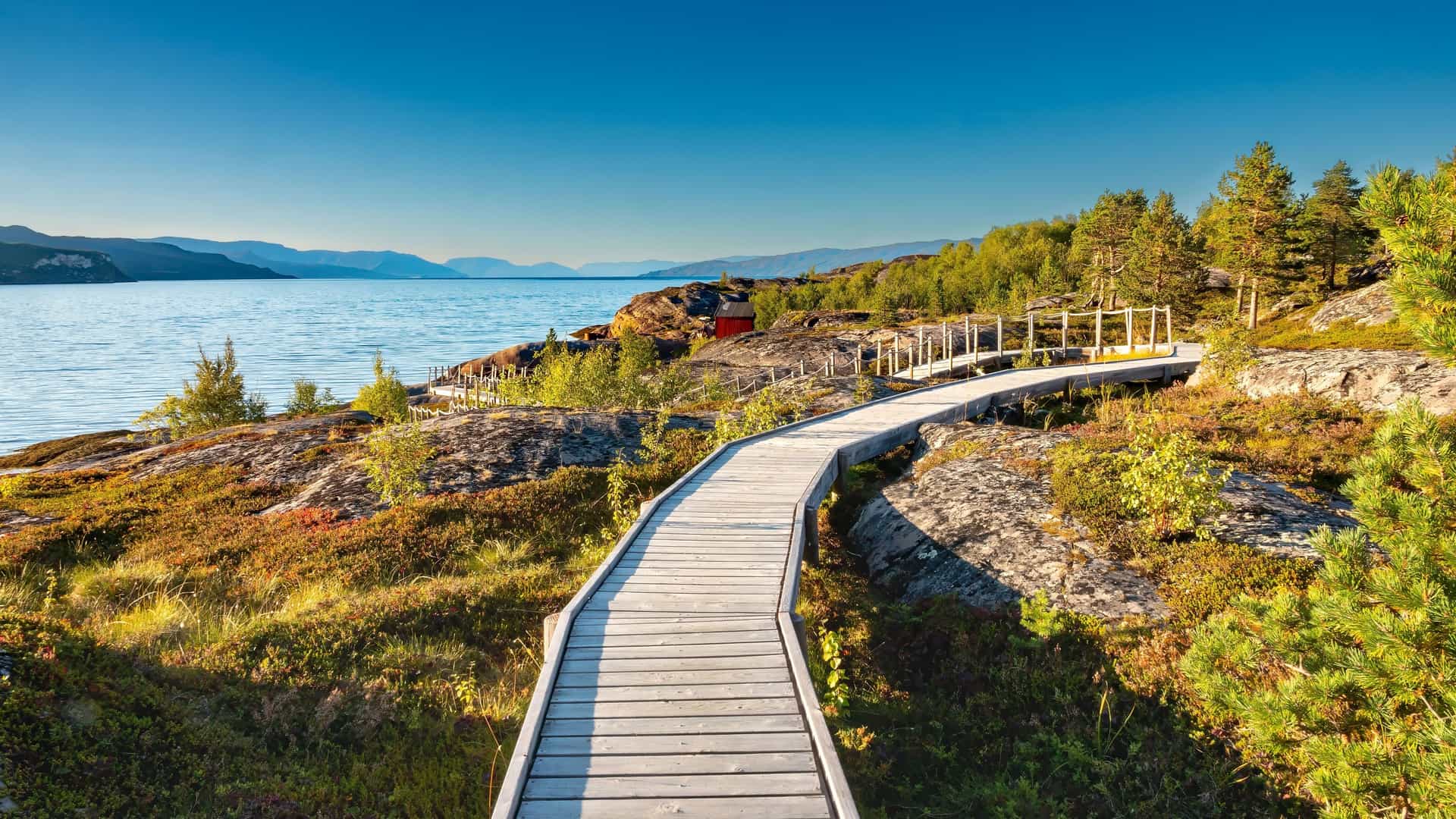 A scenic wooden boardwalk leads along the rugged shoreline of the Altafjord in Norway, with a small red house and pine trees on the shore, and blue mountains in the background.