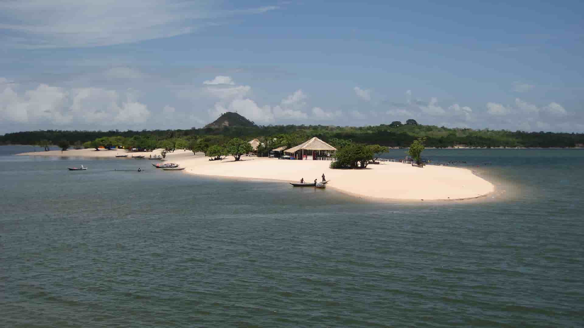 An aerial view of a sandbar in Alter do Chão, Brazil, with a few small huts and boats, surrounded by the clear, calm waters of the Tapajós River.