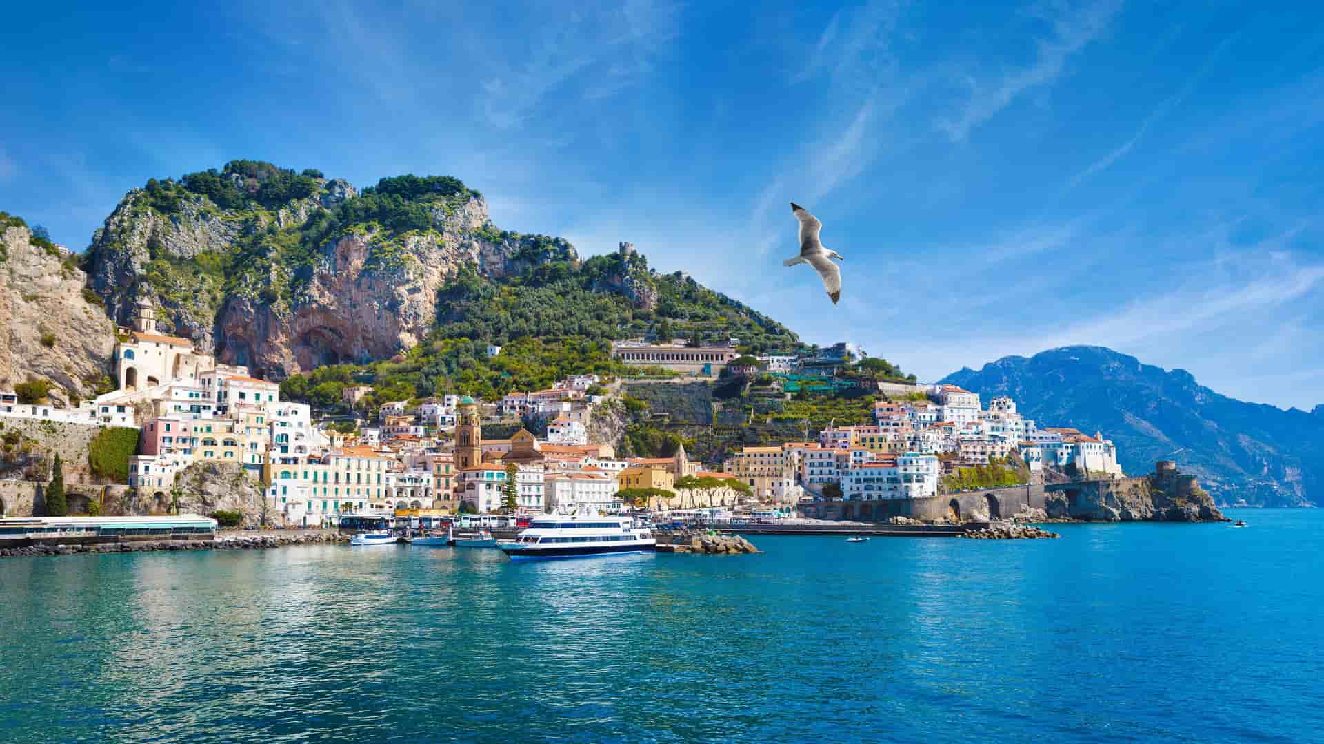 A stunning panoramic shot of the Amalfi Coast's colorful, terraced houses built into the dramatic cliffs overlooking the turquoise Mediterranean Sea, with a seagull flying overhead.