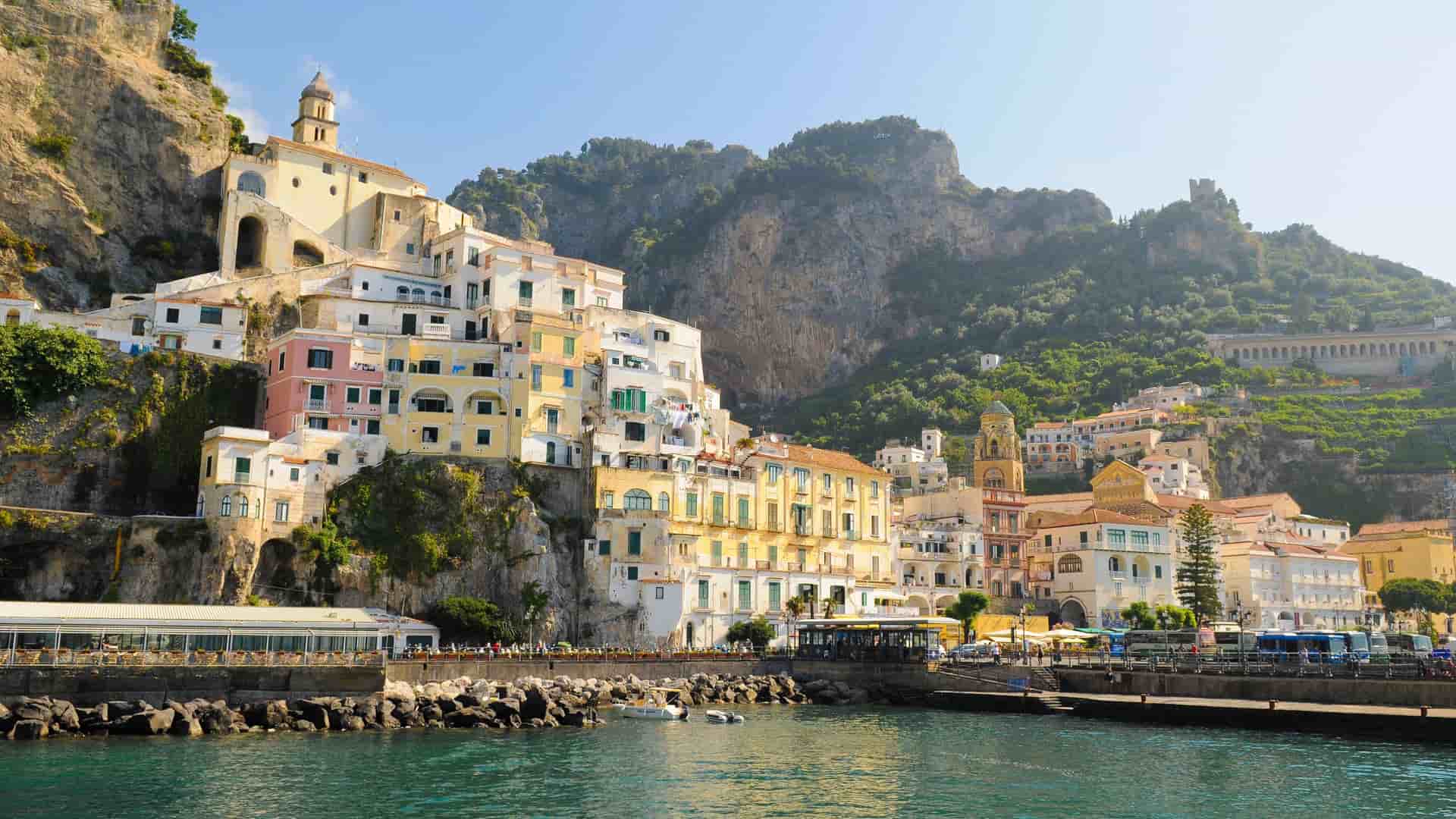 A sunny waterfront view of the colorful, cliffside village of Amalfi in Italy, with historic buildings and mountains rising from the Tyrrhenian Sea.