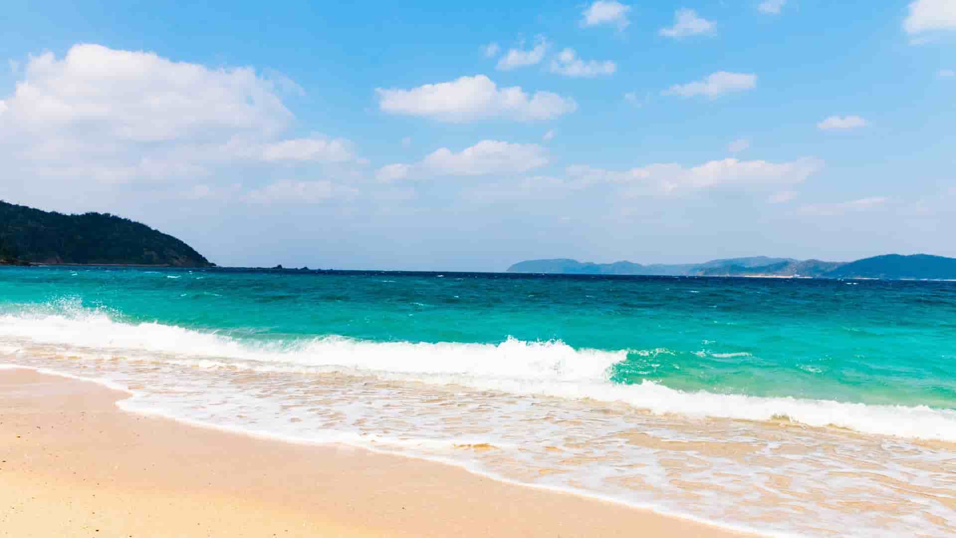 A sunny seascape of a sandy beach in Amami Ćshima, Japan, with turquoise waves crashing onto the shore and a lush, green island in the distance.
