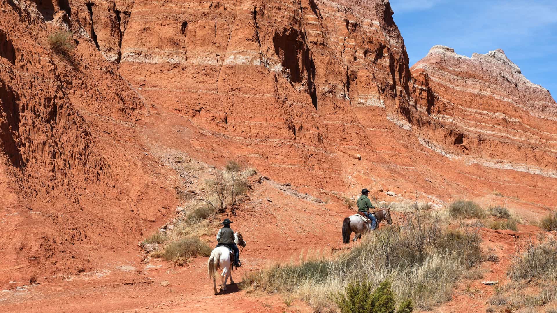Two people on horseback ride through the rugged, red-rock landscape of Palo Duro Canyon near Amarillo, Texas. The horses and riders are in the foreground, with massive, layered red and white cliffs rising dramatically behind them under a bright, clear blue sky.