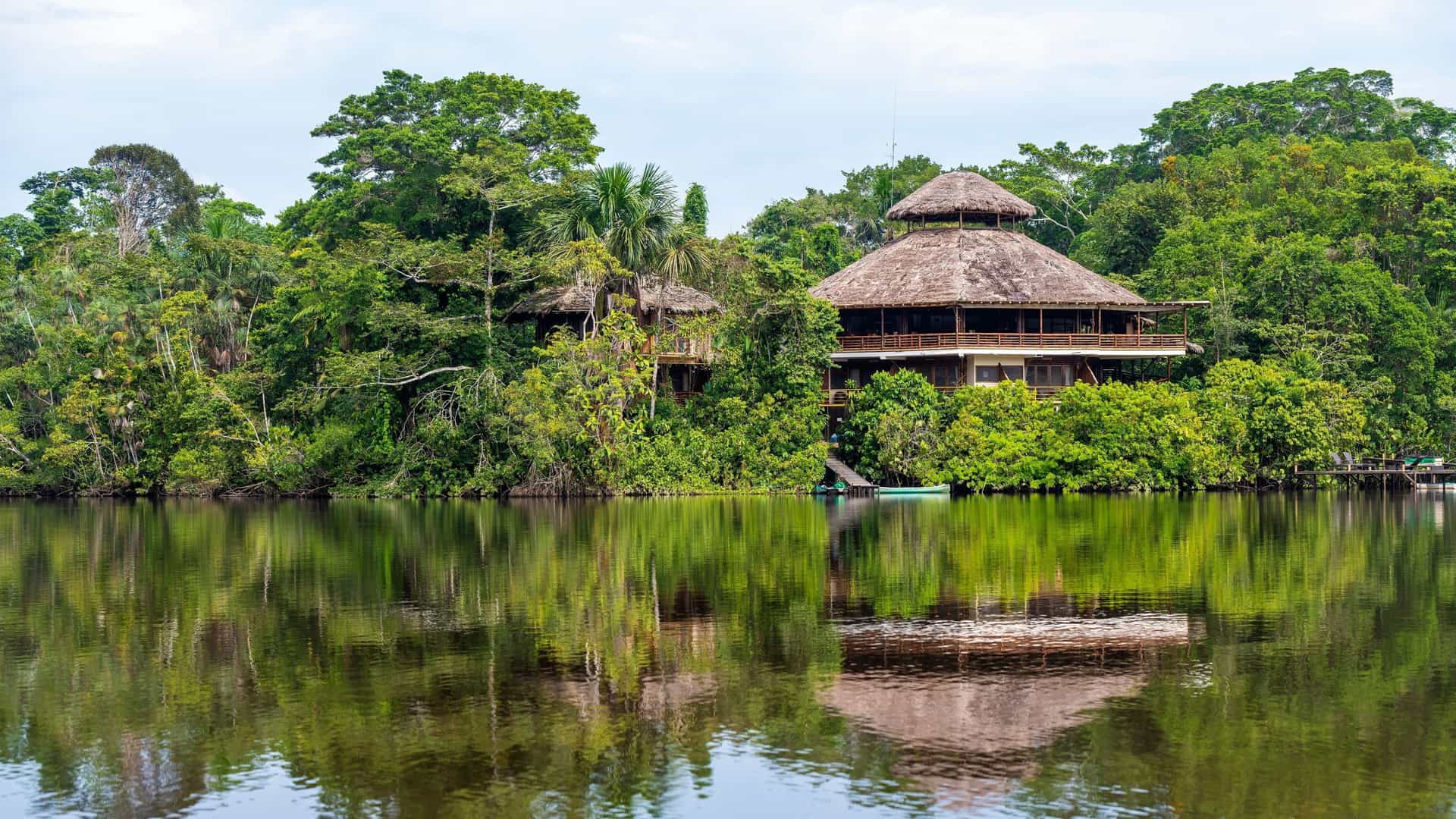 A beautiful two-story eco-lodge with a thatched roof, built on stilts and nestled within the dense, lush Amazon rainforest, with its reflection visible in the calm river water.