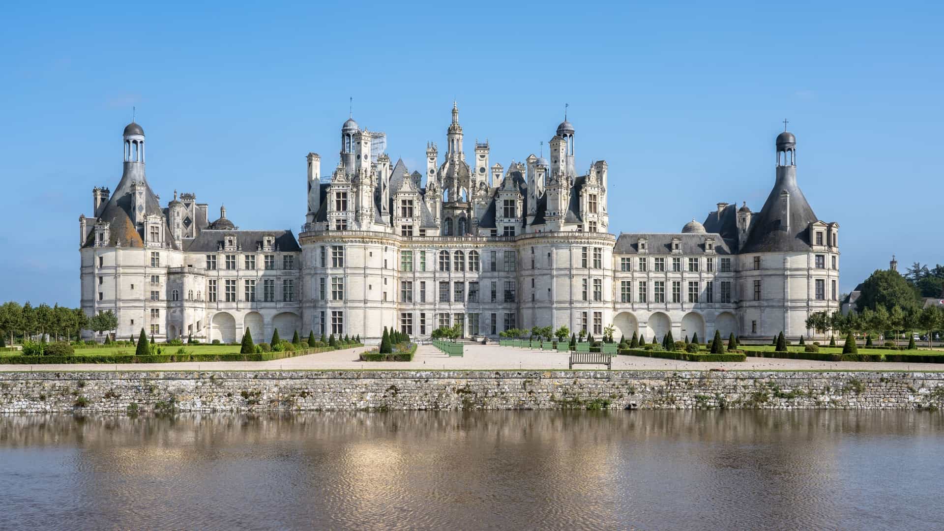 A magnificent wide-shot of the historic Château de Chambord, a Renaissance castle with ornate turrets, reflected in the still waters of the Loire River in Amboise, France.