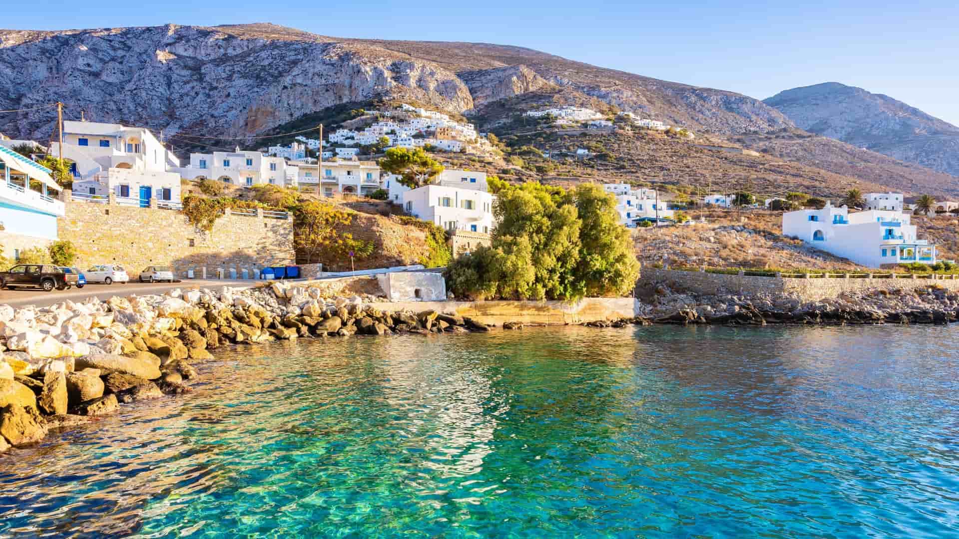"A stunning view of the whitewashed buildings of a seaside village on the Greek island of Amorgos, built on the steep hillside overlooking the clear turquoise Aegean Sea. "
