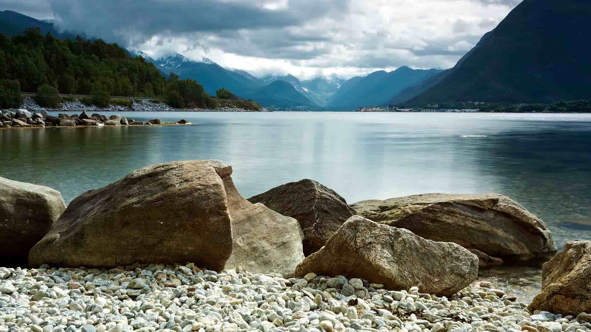 A dramatic landscape of the Romsdalsfjord in Åndalsnes, Norway, with large rocks in the foreground and majestic snow-capped mountains and a cloudy sky in the background.