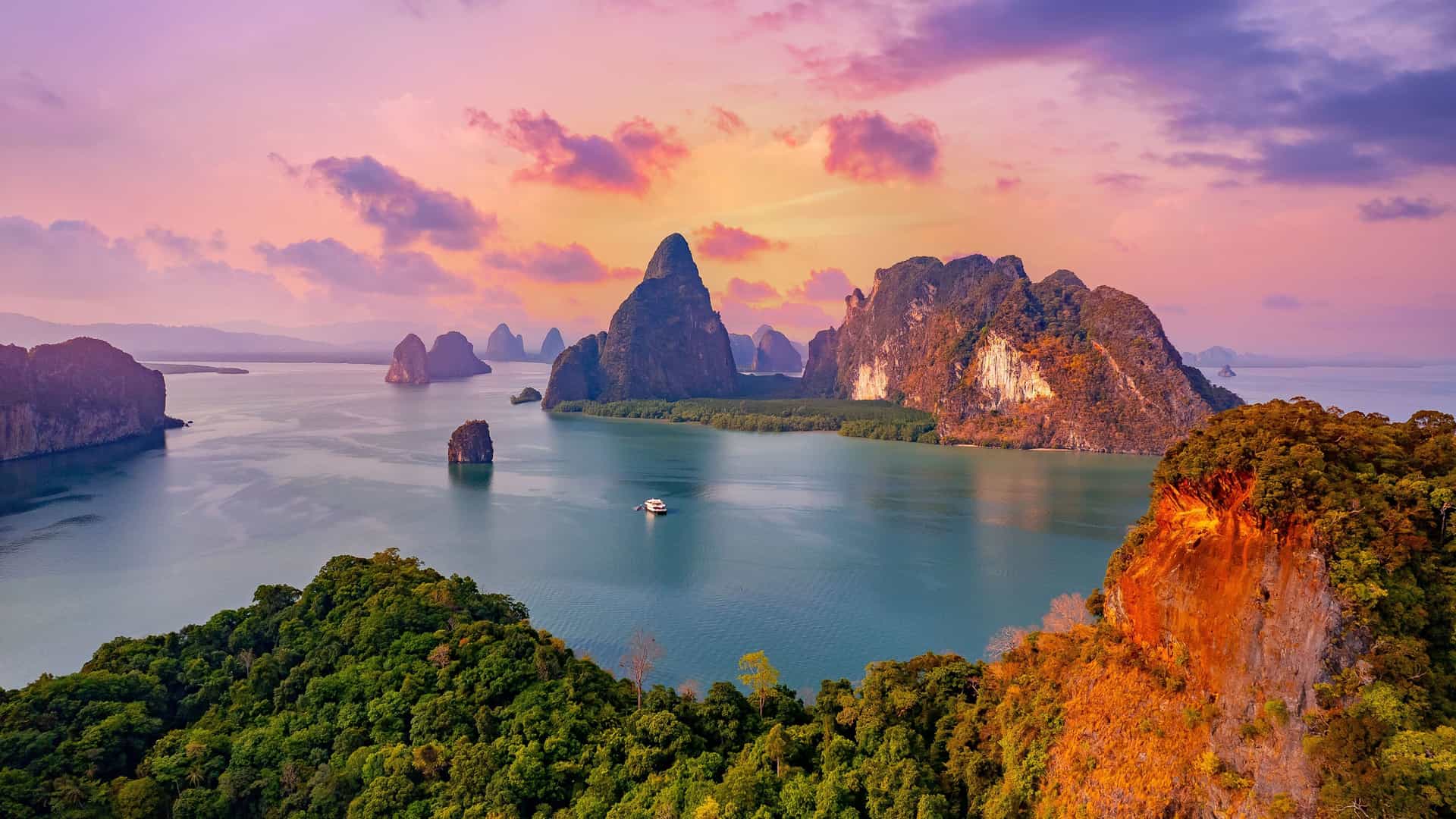 A wide-angle sunset view of the Andaman Sea's iconic limestone cliffs and rock formations rising from the serene waters, with a boat sailing in the foreground.