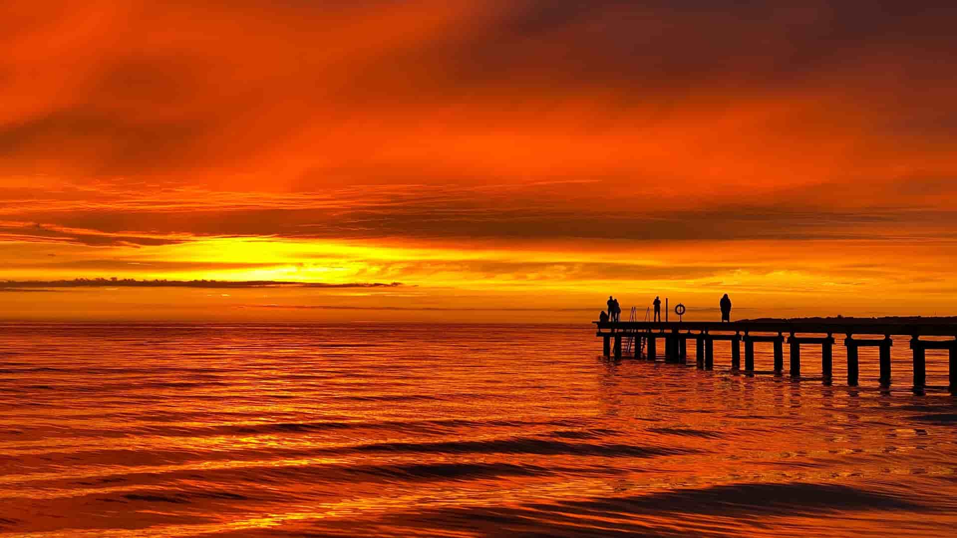 A beautiful sunset photograph from Helsingborg, Sweden, showing silhouettes of people on a pier against a brilliant orange and red sky reflected on the still water.