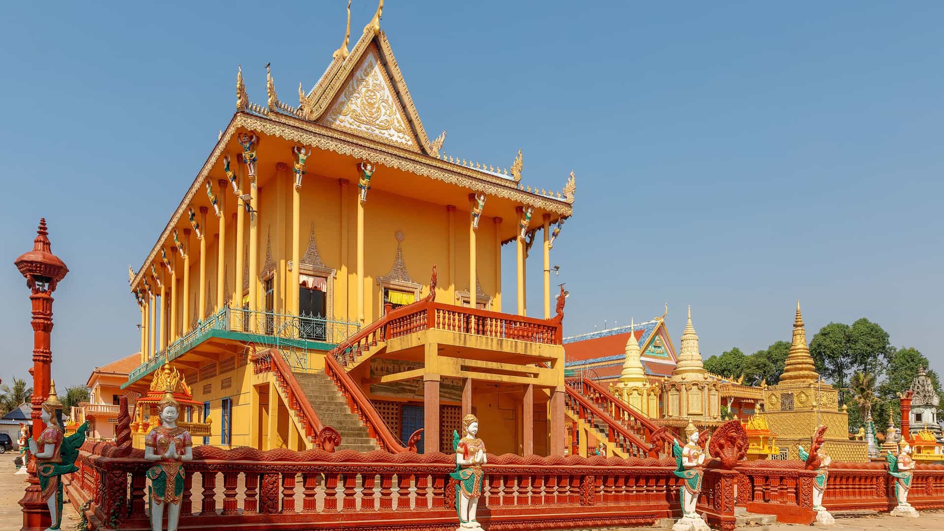 A sunny wide shot of the vibrant yellow Wat Kbal Toan temple, a Cambodian landmark in the village of Angkor Ban, featuring ornate decorations and a large staircase.