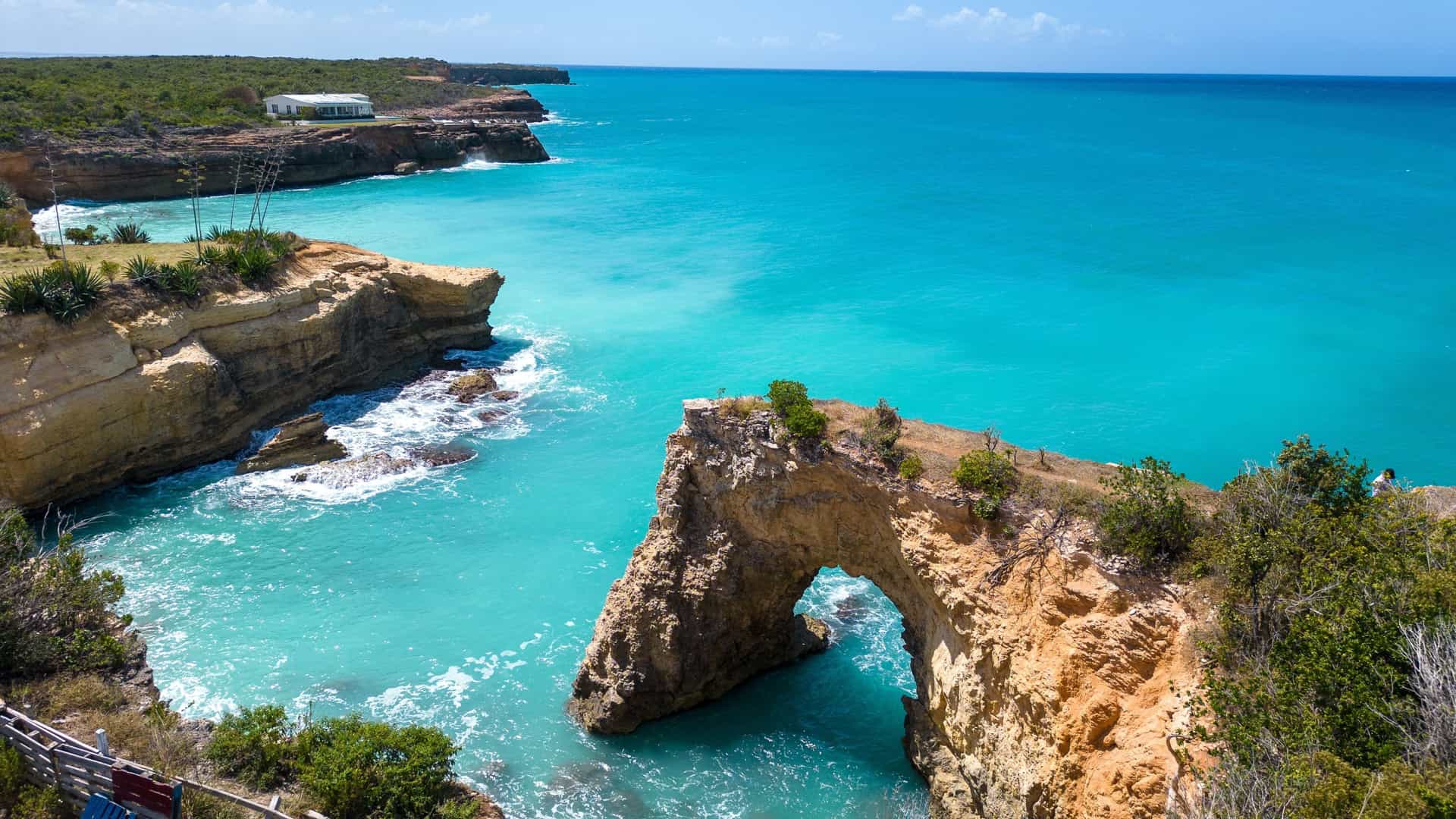A dramatic coastal scene in Anguilla featuring a natural rock arch rising from turquoise water, surrounded by rugged cliffs with sparse vegetation and a white building perched above the shoreline.