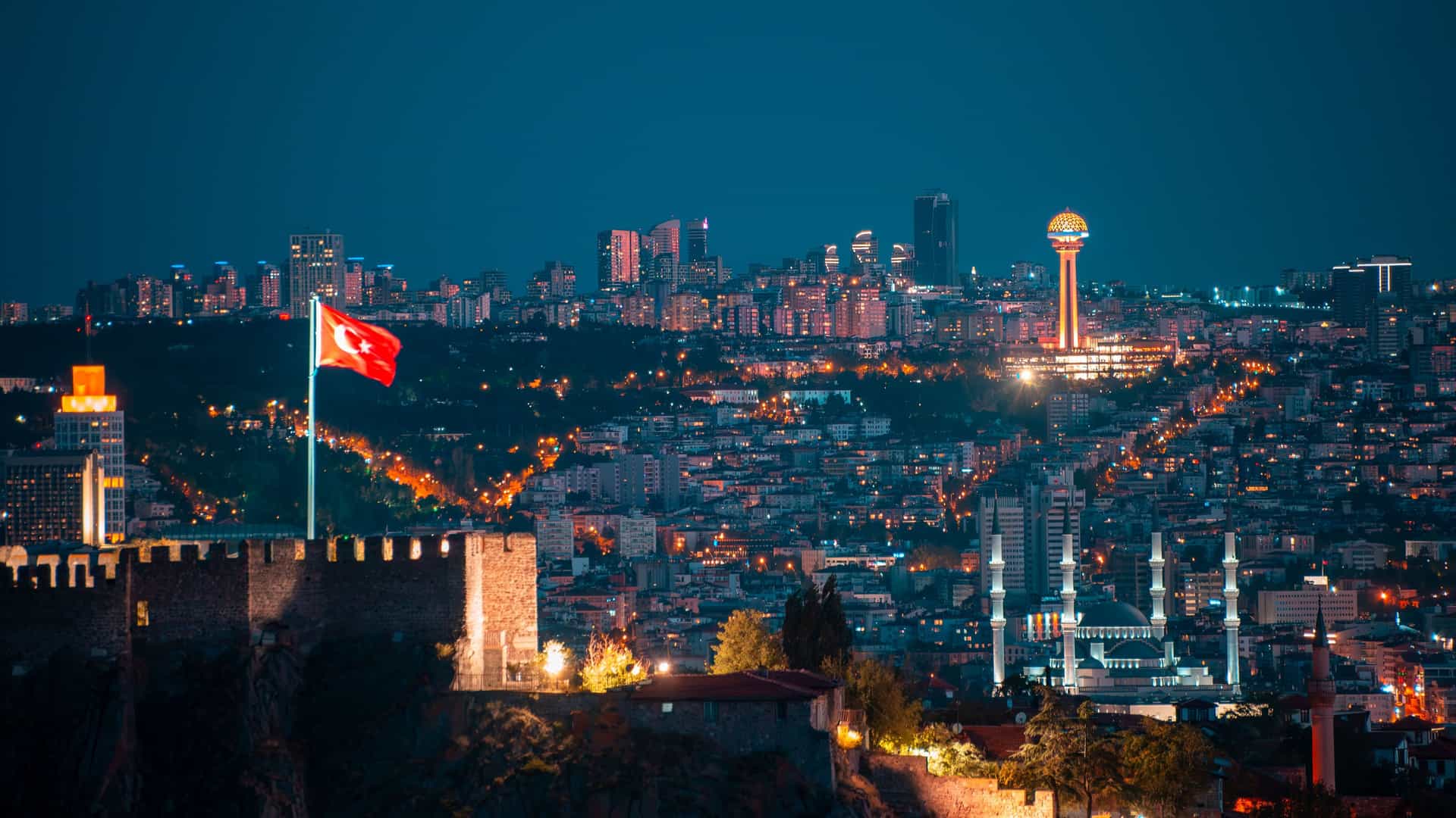 A panoramic night view of the city of Ankara, Turkey. In the foreground, the ancient stone walls of Ankara Castle are visible, with a large Turkish flag flying high. The sprawling cityscape in the background is a brilliant tapestry of lights, highlighted by the brightly lit Atakule tower and a large mosque.