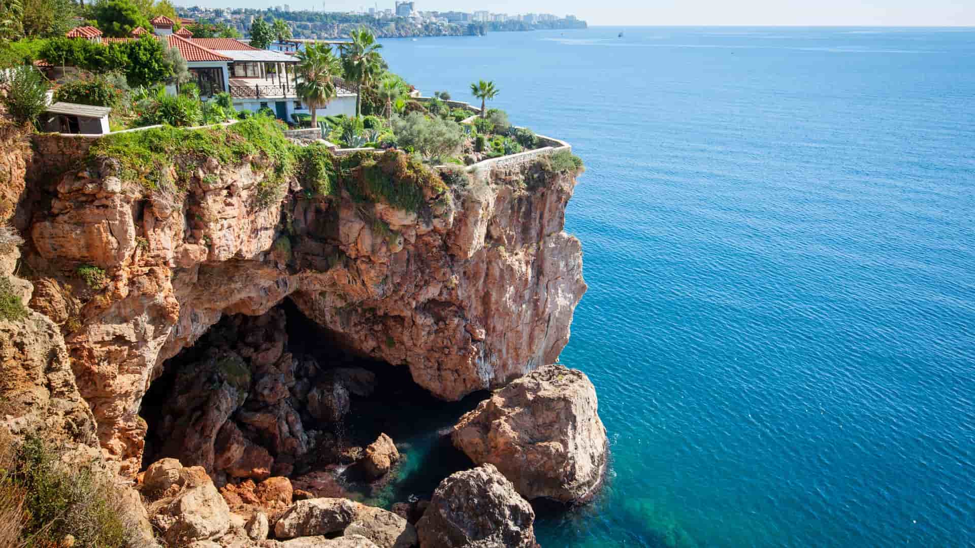 A sunny cliffside view of the clear blue Mediterranean Sea in Antalya, Turkey, with a beautiful house and palm trees perched on the edge of the rocky cliffs.