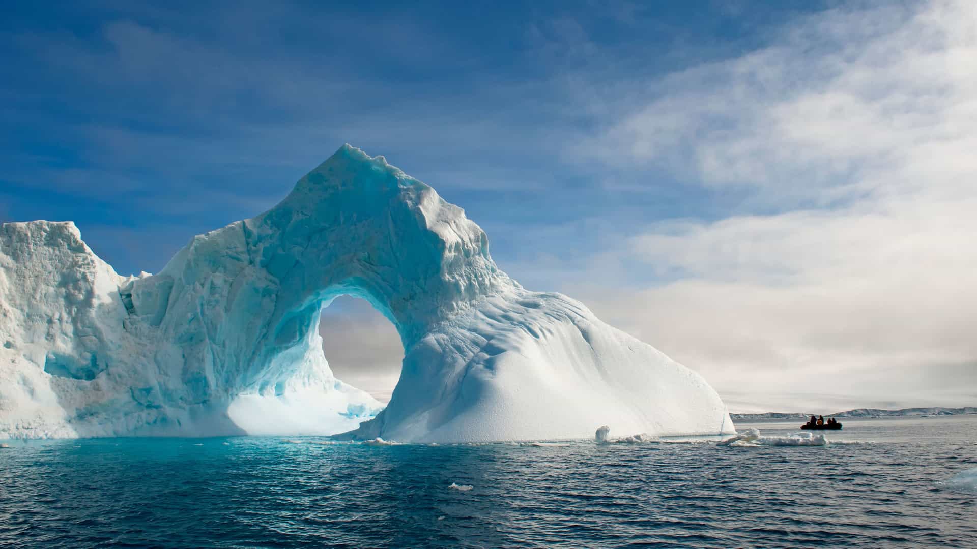 A zodiac boat carrying passengers cruises near a massive, naturally formed archway in an iceberg, showcasing the stunning scenery of the Antarctic Sound.