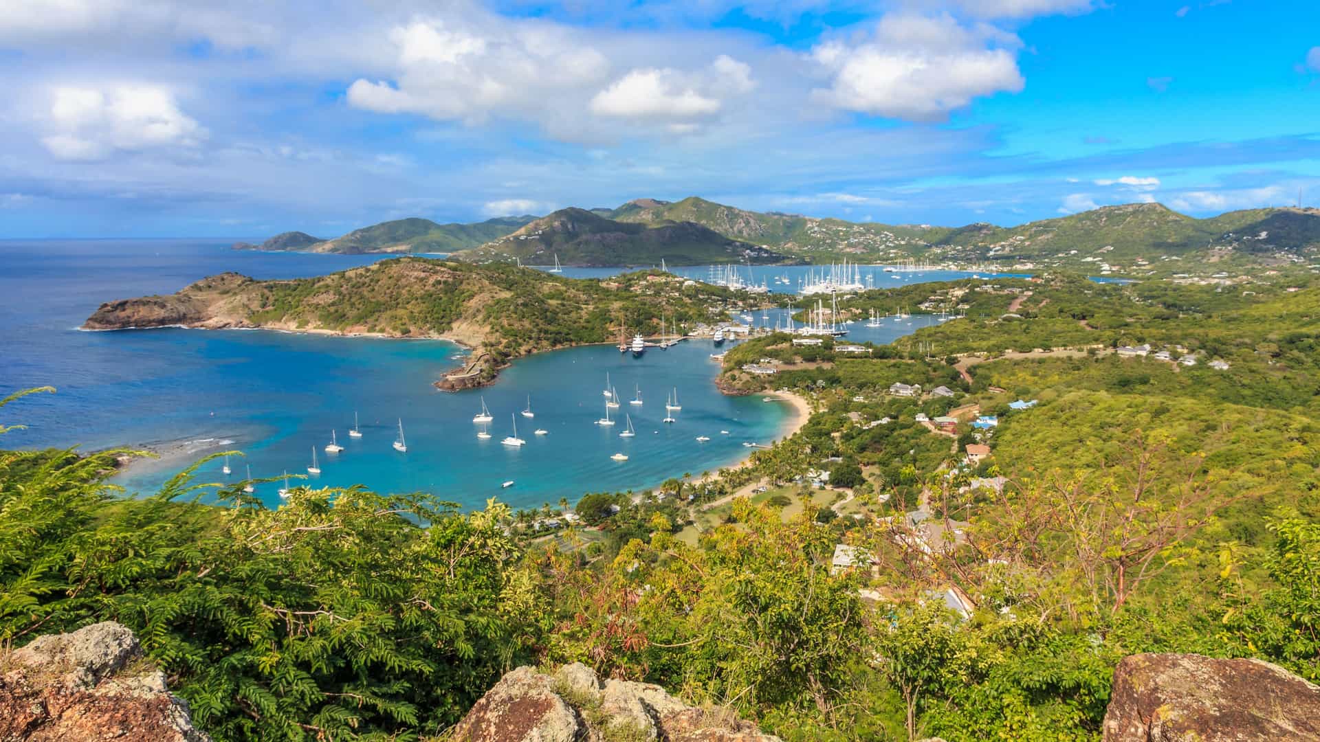 A panoramic aerial shot of the beautiful English Harbour in Antigua, showing the turquoise waters of the Caribbean Sea, dotted with sailboats and surrounded by lush green hills.