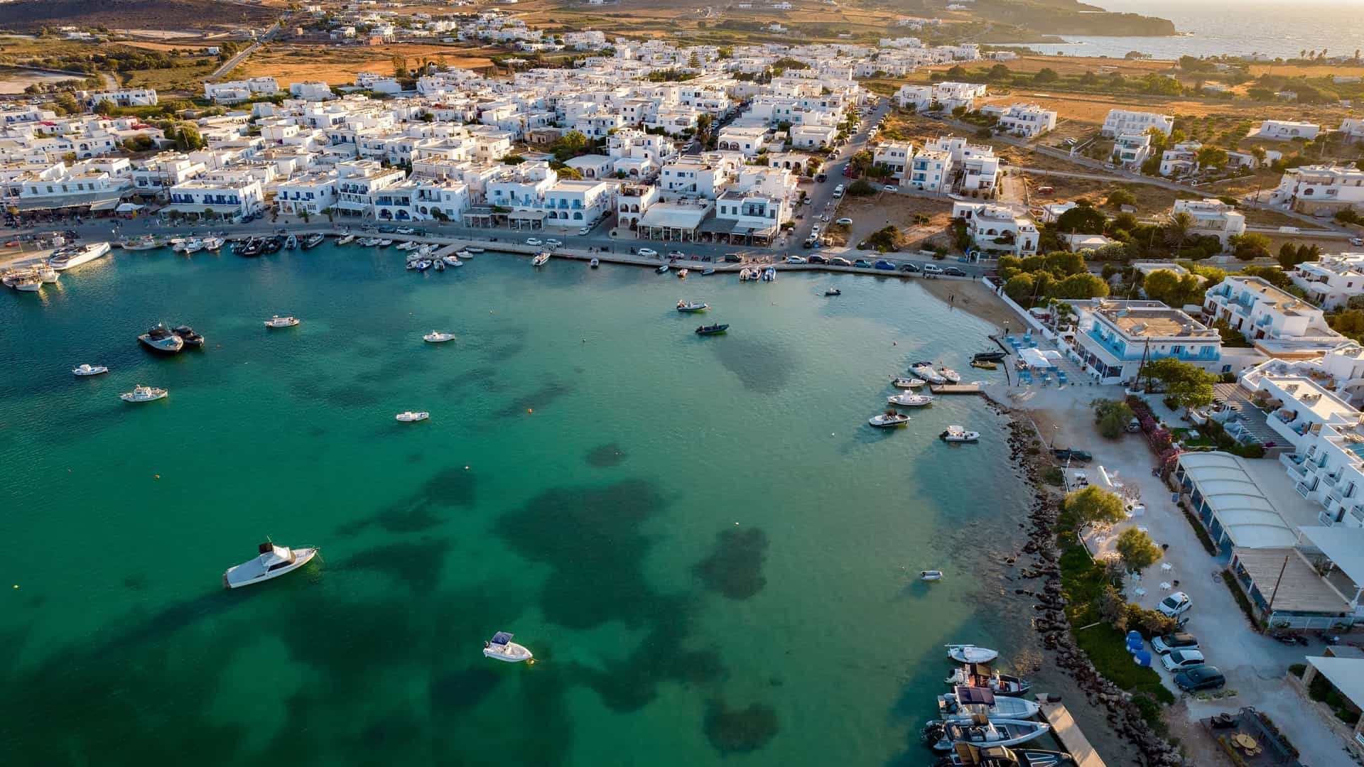 An aerial view of the coastal town of Antiparos, Greece, with its whitewashed Cycladic buildings clustered around a turquoise-colored harbor. Numerous small boats are moored in the clear water, which transitions to a deeper blue further out. The buildings of the town extend up into the dry, golden hills under a bright, sunny sky.