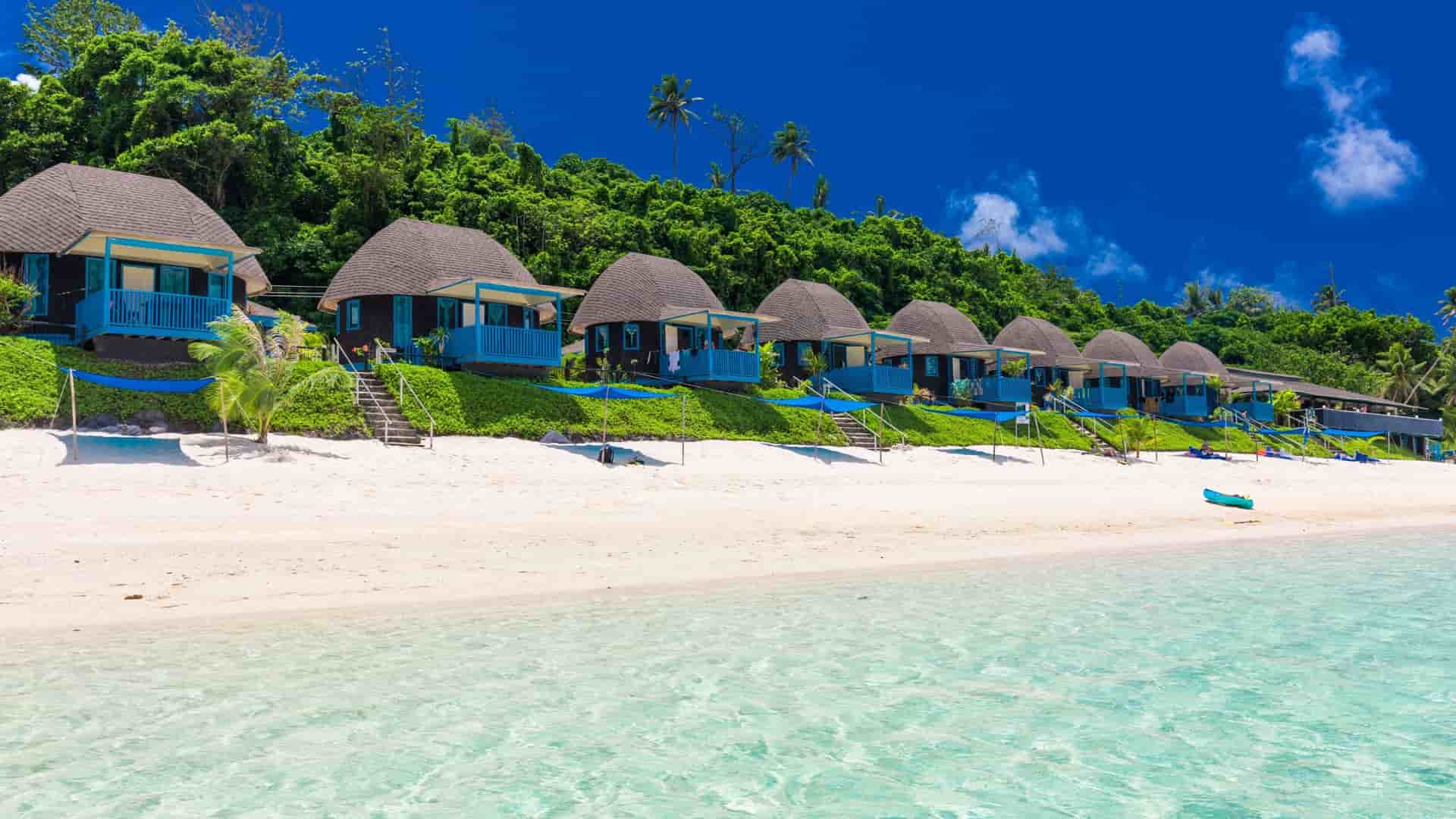 A scenic shot of a line of small, blue, thatched-roof beach bungalows on a pristine white sand beach in Apia, Samoa, with a clear turquoise ocean and a lush green hill in the background.
