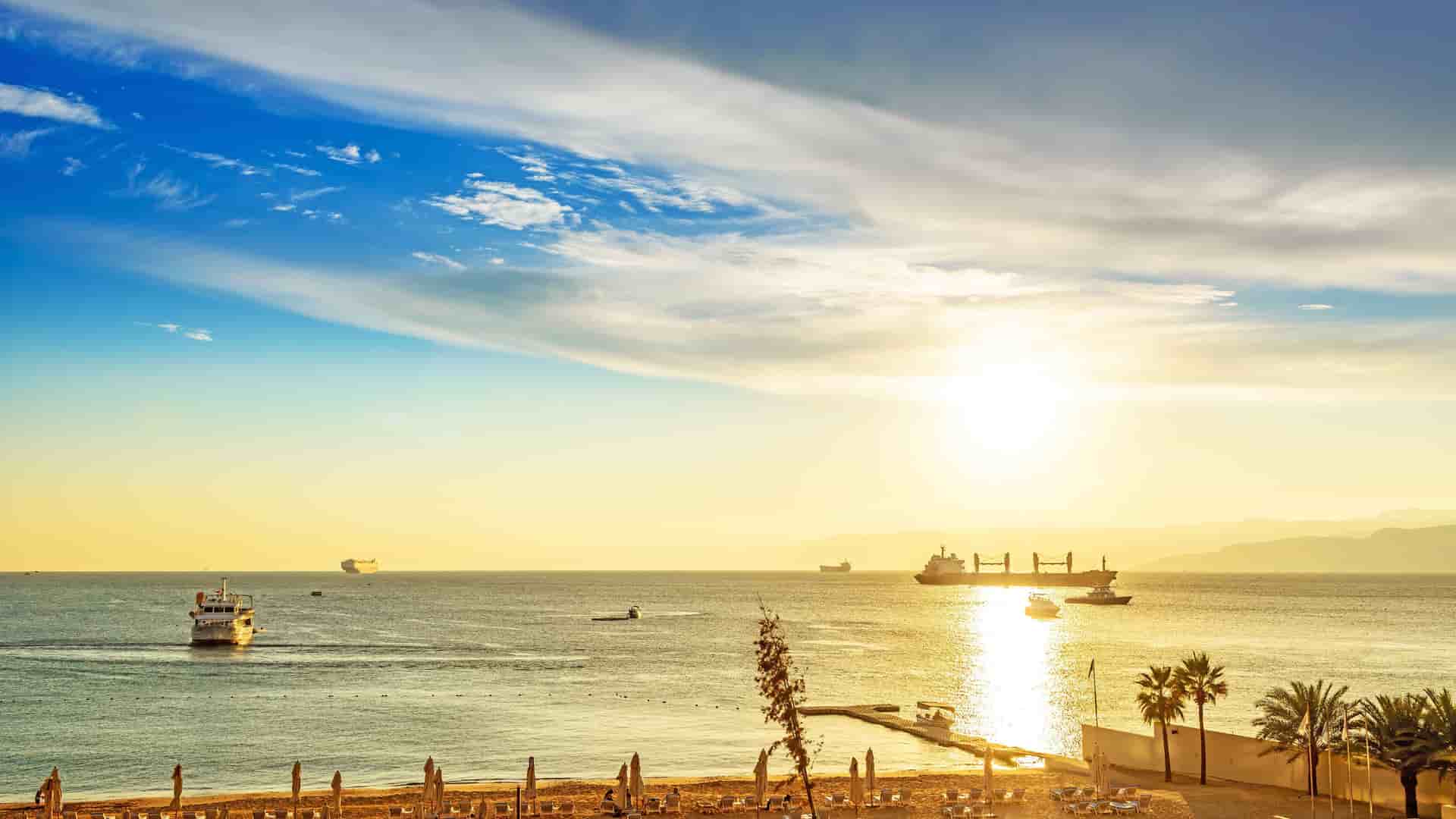 A scenic sunset view over the Gulf of Aqaba, with the sun setting behind the mountains, casting a golden glow on the water and a beach with small boats and large ships on the horizon.