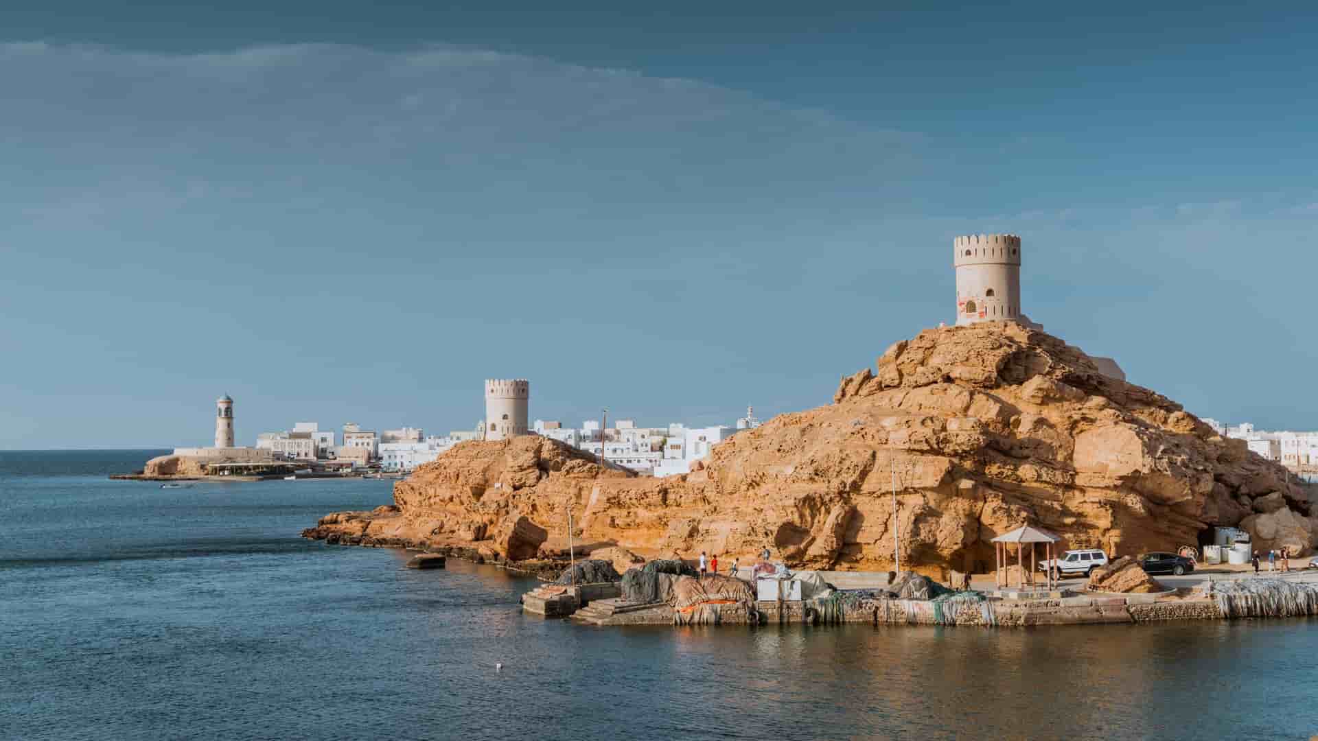 A panoramic shot of Sur, Oman, showing its historic watchtowers on rocky cliffs overlooking the Arabian Sea, with the city's whitewashed buildings in the distance.