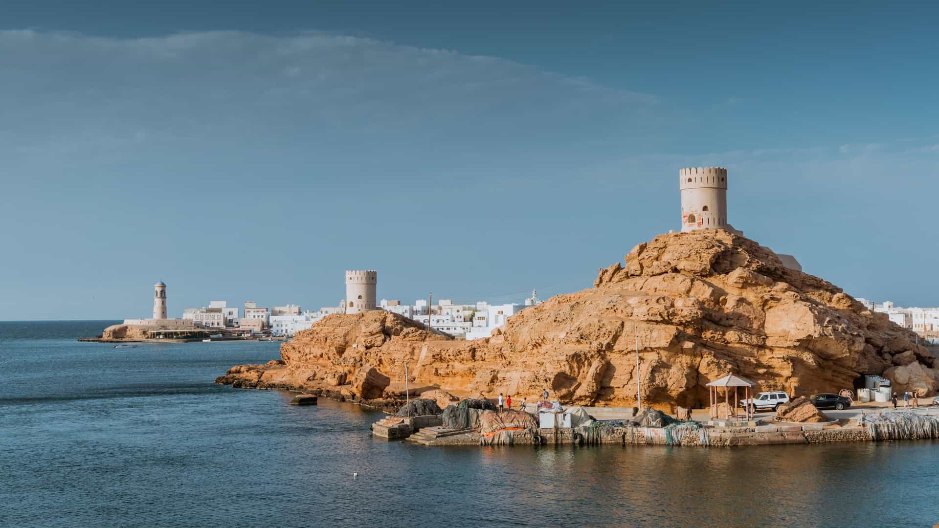 A panoramic shot of Sur, Oman, showing its historic watchtowers on rocky cliffs overlooking the Arabian Sea, with the city's whitewashed buildings in the distance.