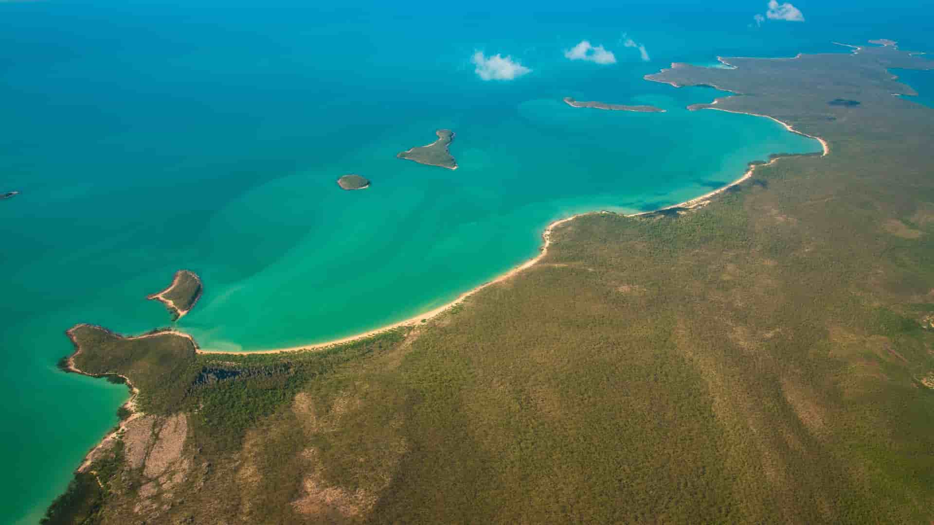 An aerial view of the stunning Arafura Sea coastline, showcasing a pristine sandy beach curving around a bay with turquoise waters and lush green land in the foreground.