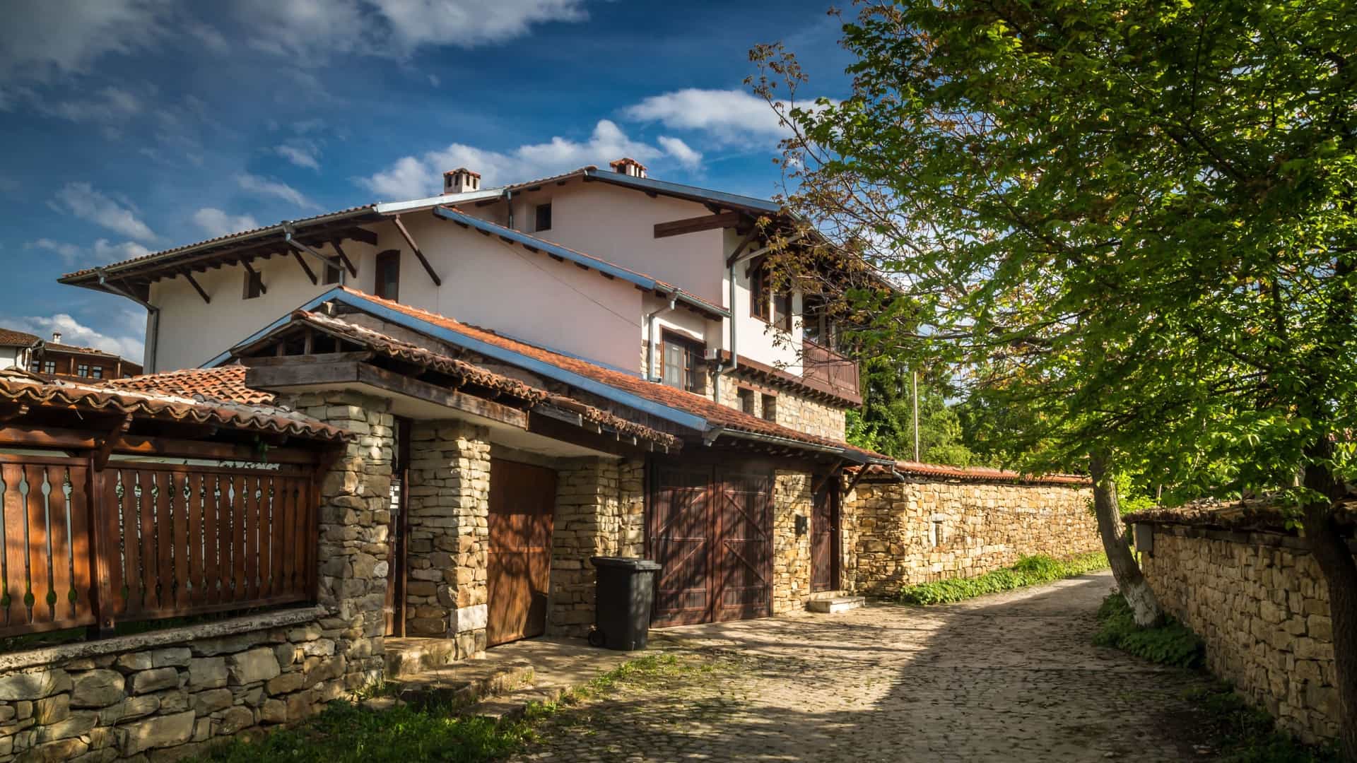 A sunny, wide view of a traditional Arbanassi-style house in Bulgaria, featuring a stone wall, cobblestone path, and lush green trees.