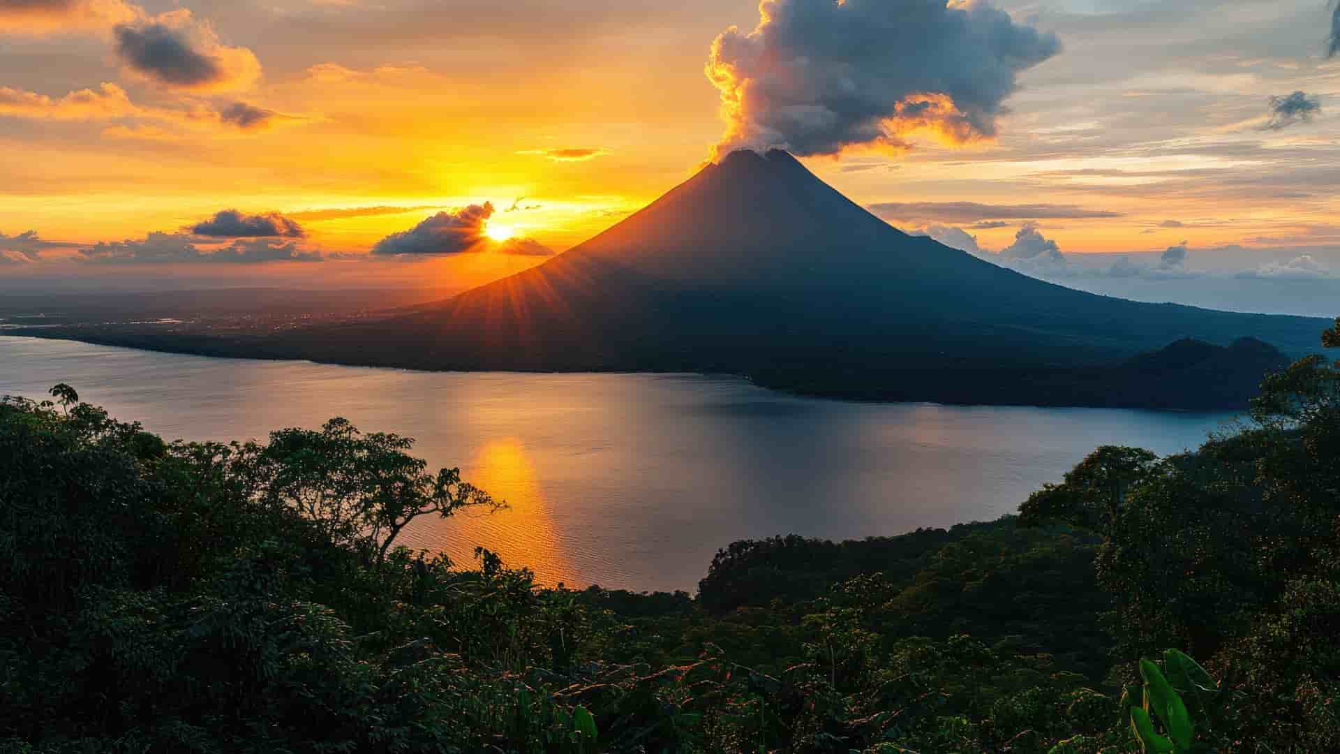 A dramatic sunset over Lake Arenal in Costa Rica, with a majestic, conical Arenal Volcano emitting a plume of smoke and fiery clouds into the orange sky.