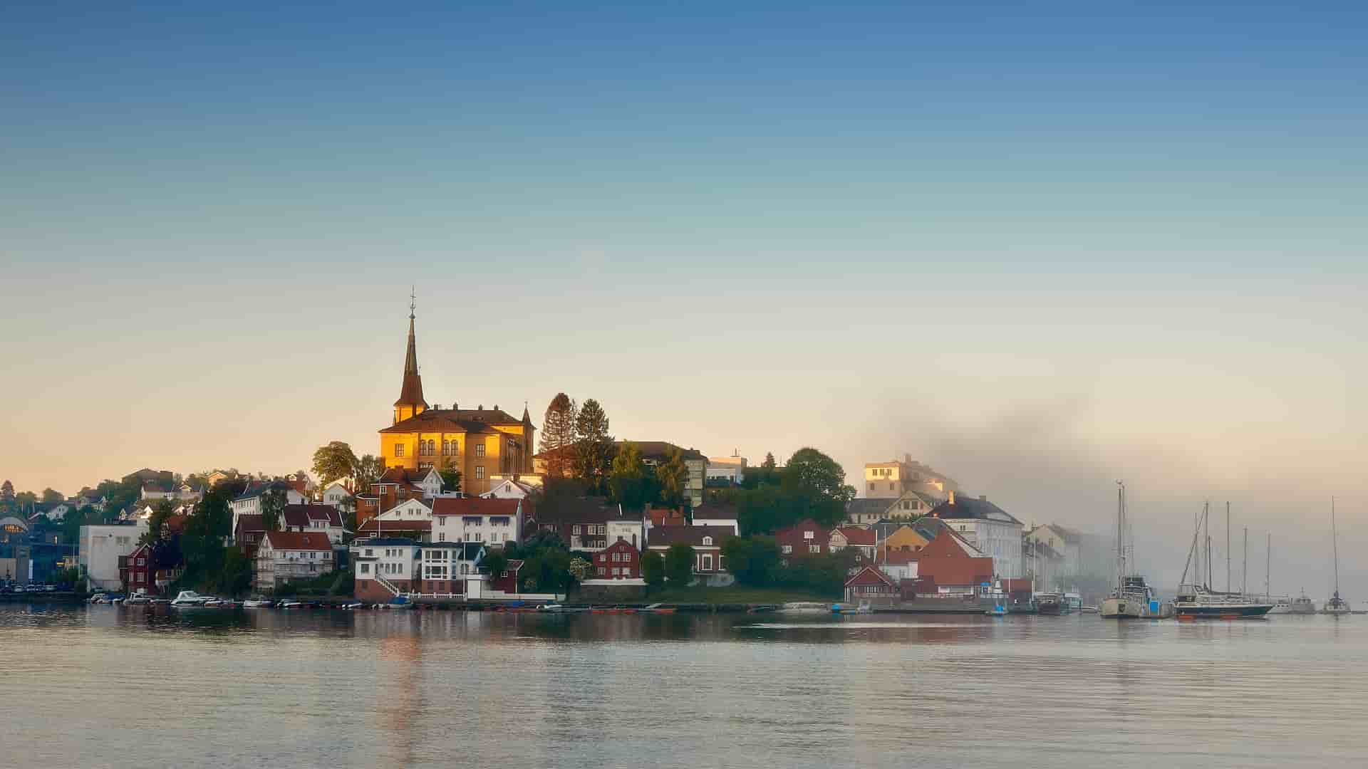 A stunning sunrise shot of the historic town of Arendal, Norway, with its church steeple and colorful buildings silhouetted against a golden sky and reflected in the calm fjord waters.