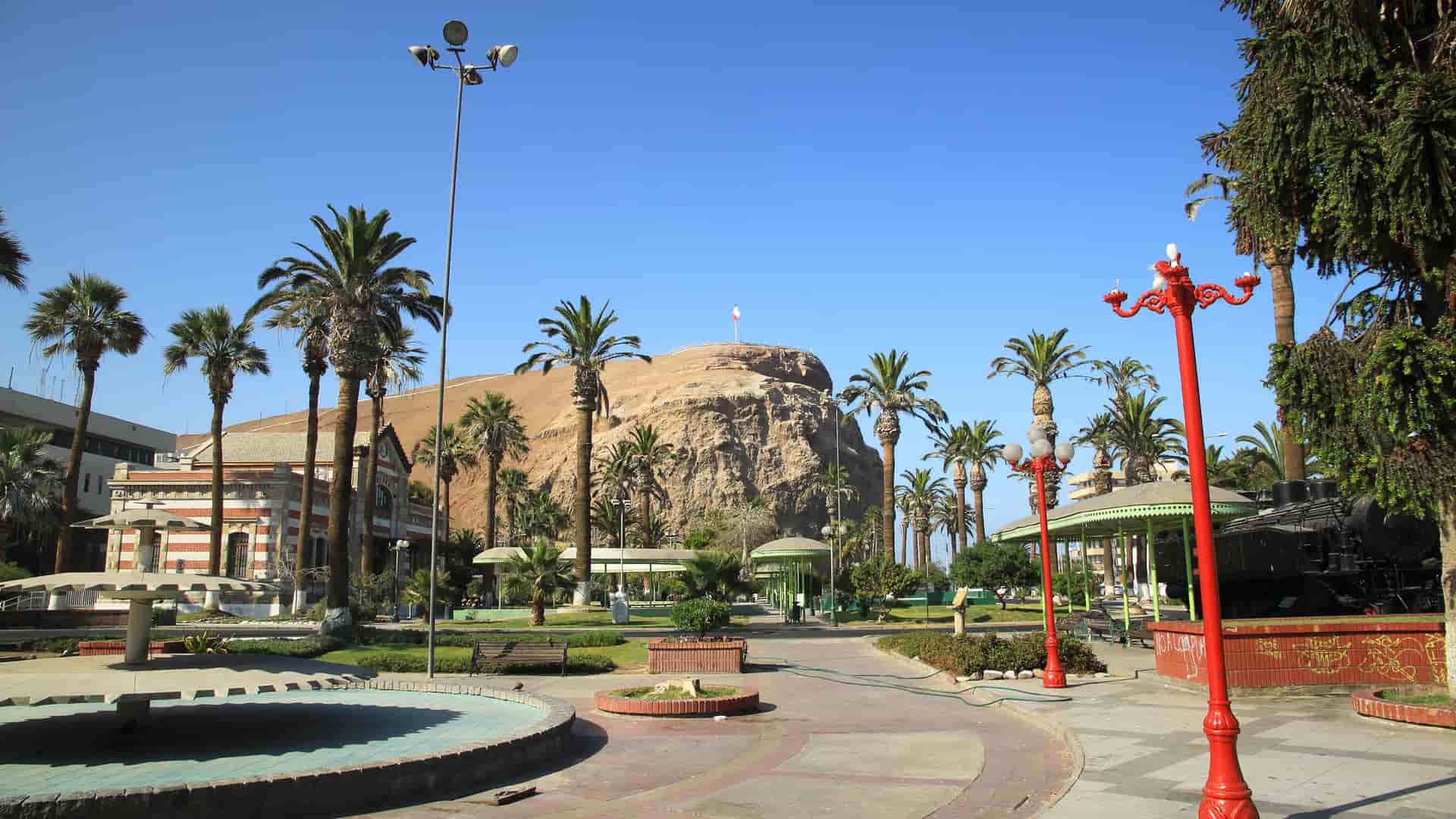 A sunny, palm tree-lined park in Arica, Chile, with historic buildings and the prominent El Morro de Arica hill visible in the background.