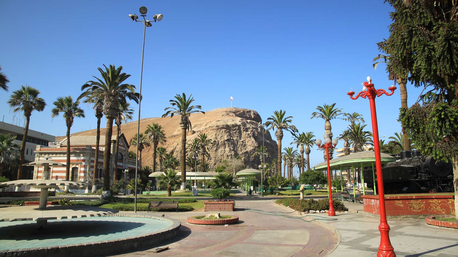 A sunny, palm tree-lined park in Arica, Chile, with historic buildings and the prominent El Morro de Arica hill visible in the background.