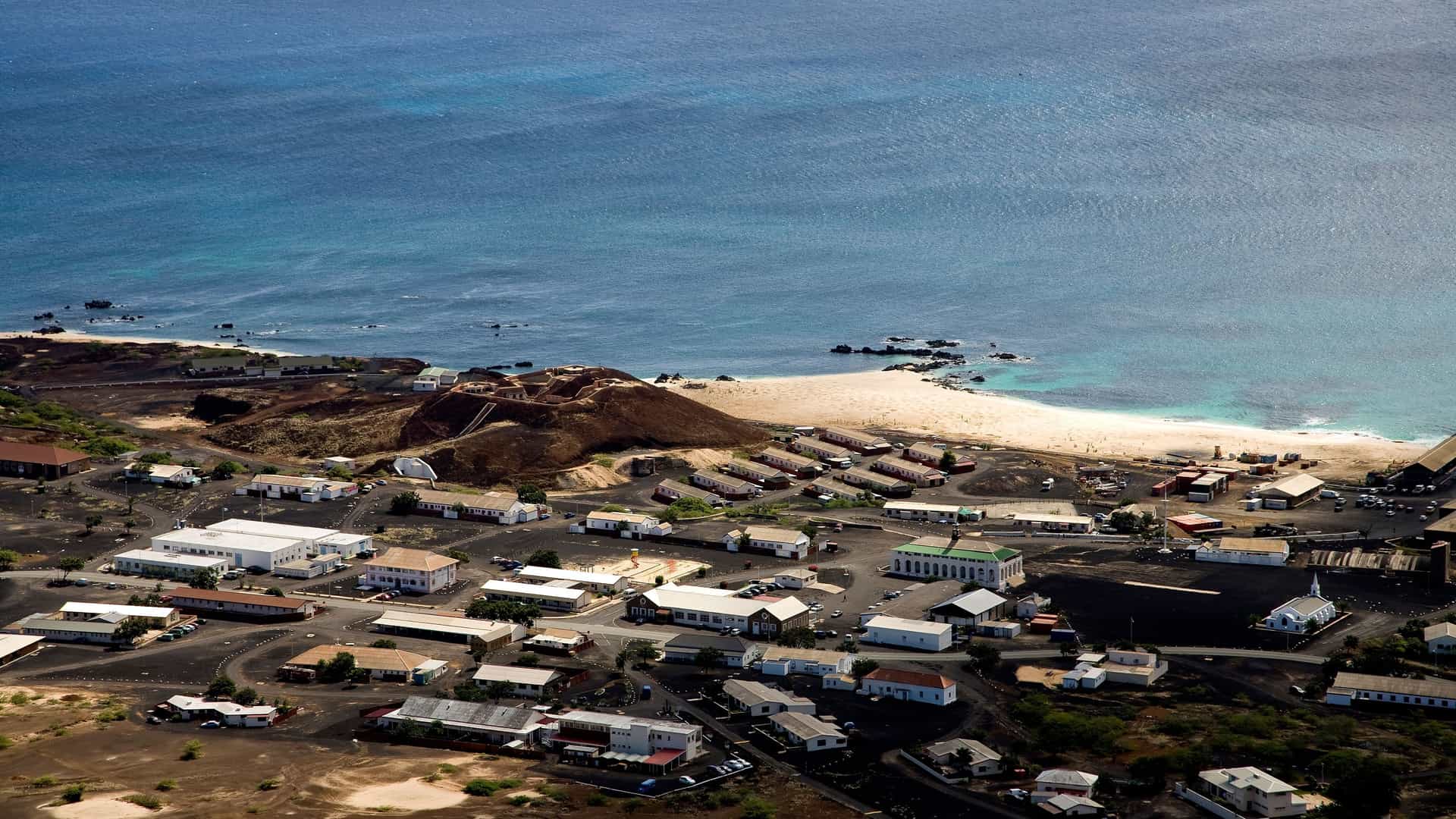 An aerial view of Georgetown on Ascension Island, showcasing the town's buildings nestled against a volcanic landscape and a sandy beach by the Atlantic Ocean.