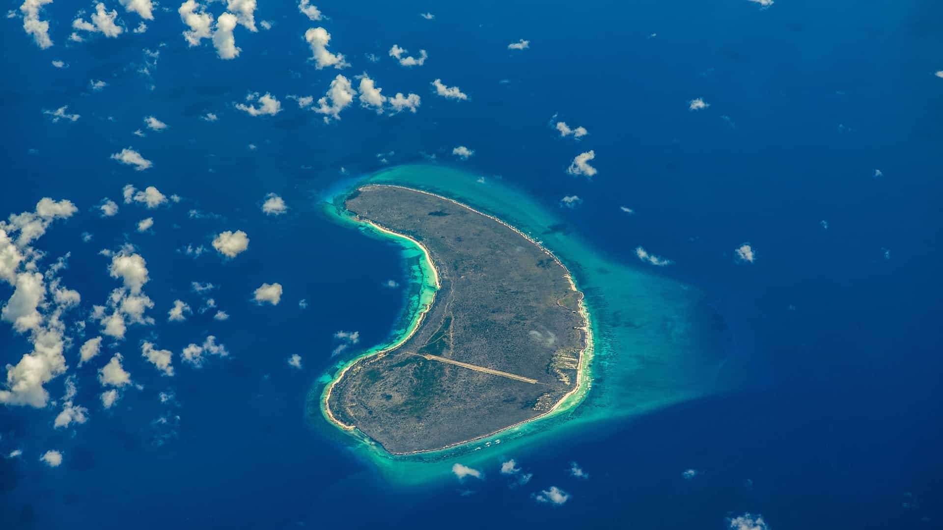 An aerial view of Assumption Island in the Seychelles, showing a small, crescent-shaped landmass with a runway surrounded by shallow turquoise water and the deep blue Indian Ocean.