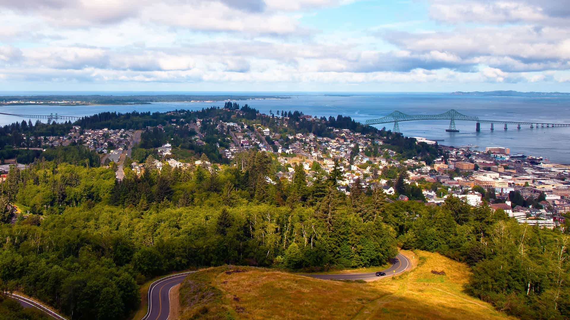 An aerial view of Astoria, Oregon, showcasing a winding road, a lush green hill, the city's residential area, and the Astoria-Megler Bridge crossing the Columbia River.