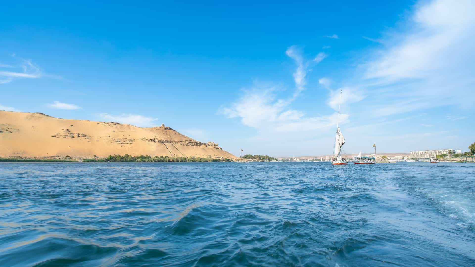 A scenic stretch of the Nile River in Aswan High Dam with a sailboat gliding across blue water, a large sandy hill topped by a small structure on one side, and modern buildings with additional boats lining the opposite riverbank.