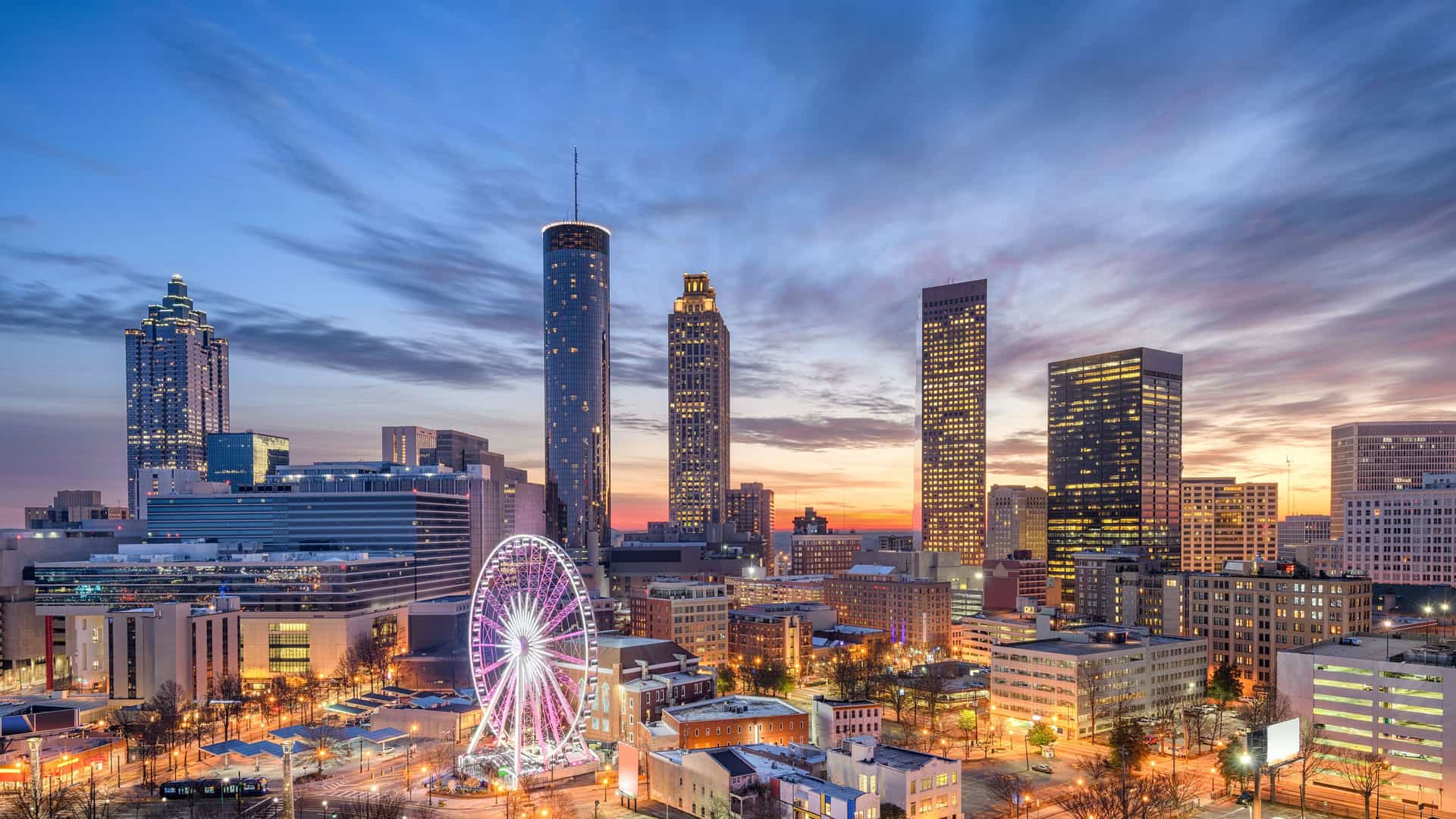 A panoramic view of the downtown Atlanta skyline at dusk, with the SkyView Atlanta Ferris wheel glowing with colorful lights in the foreground. Tall, modern skyscrapers rise into a dramatic sky painted with hues of blue, pink, and orange, as the city's lights begin to illuminate.