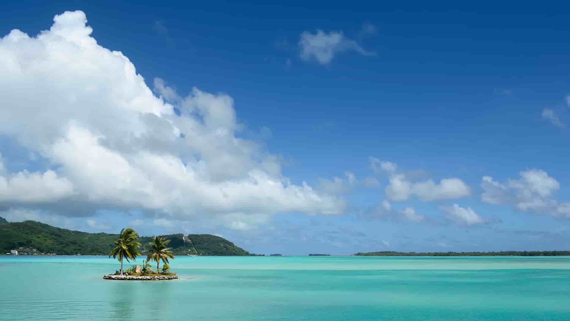 A breathtaking view of Atuona, French Polynesia, featuring a small islet with two palm trees surrounded by clear turquoise water, with lush green hills and a bright blue sky with large fluffy clouds.