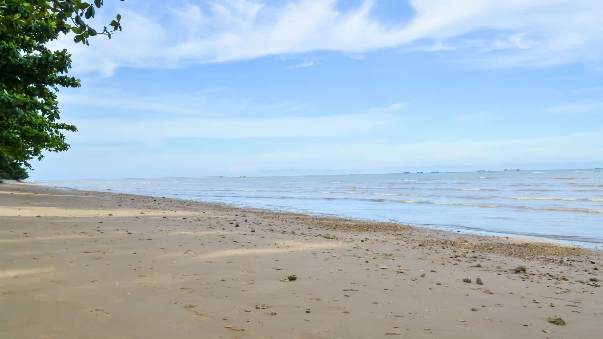 A serene view of a sandy beach in the Auri Islands, Indonesia. The quiet shoreline has light-colored sand and small pebbles that meet the calm blue sea, while a few ships are visible on the horizon under a bright sky with scattered clouds.